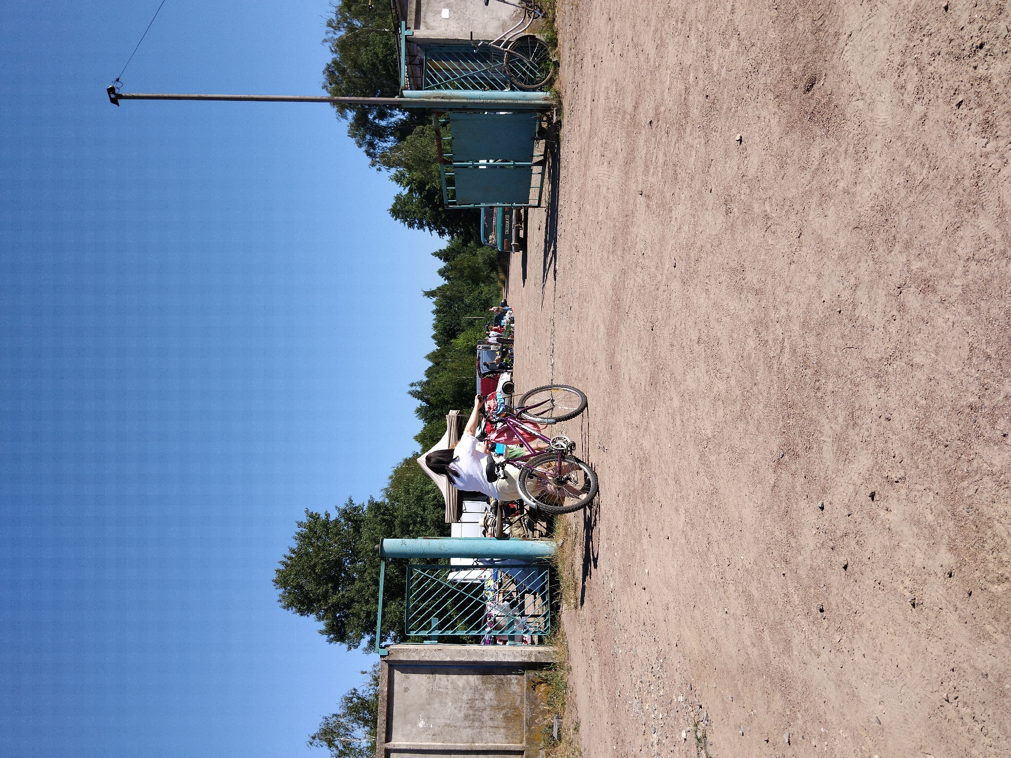 The image depicts a scene at an outdoor market or event with a young woman on a pink bicycle in the foreground. She is wearing a white top and light-colored shorts, with her back to the camera, facing towards an open gate. The bicycle is positioned on a dirt path. Behind her, through the green metal gate, appears to be a bustling market with tents, tables, and various goods displayed. Several vehicles, including a green car with a license plate that reads "M 18 49 BH", are visible in the background, along with other people browsing the stalls. The environment is outdoors, with a clear blue sky and trees lining the perimeter of the market area. A tall pole with some electrical equipment is situated to the right of the frame. The overall setting suggests a sunny day at a local marketplace in Irshansk, Ukraine.