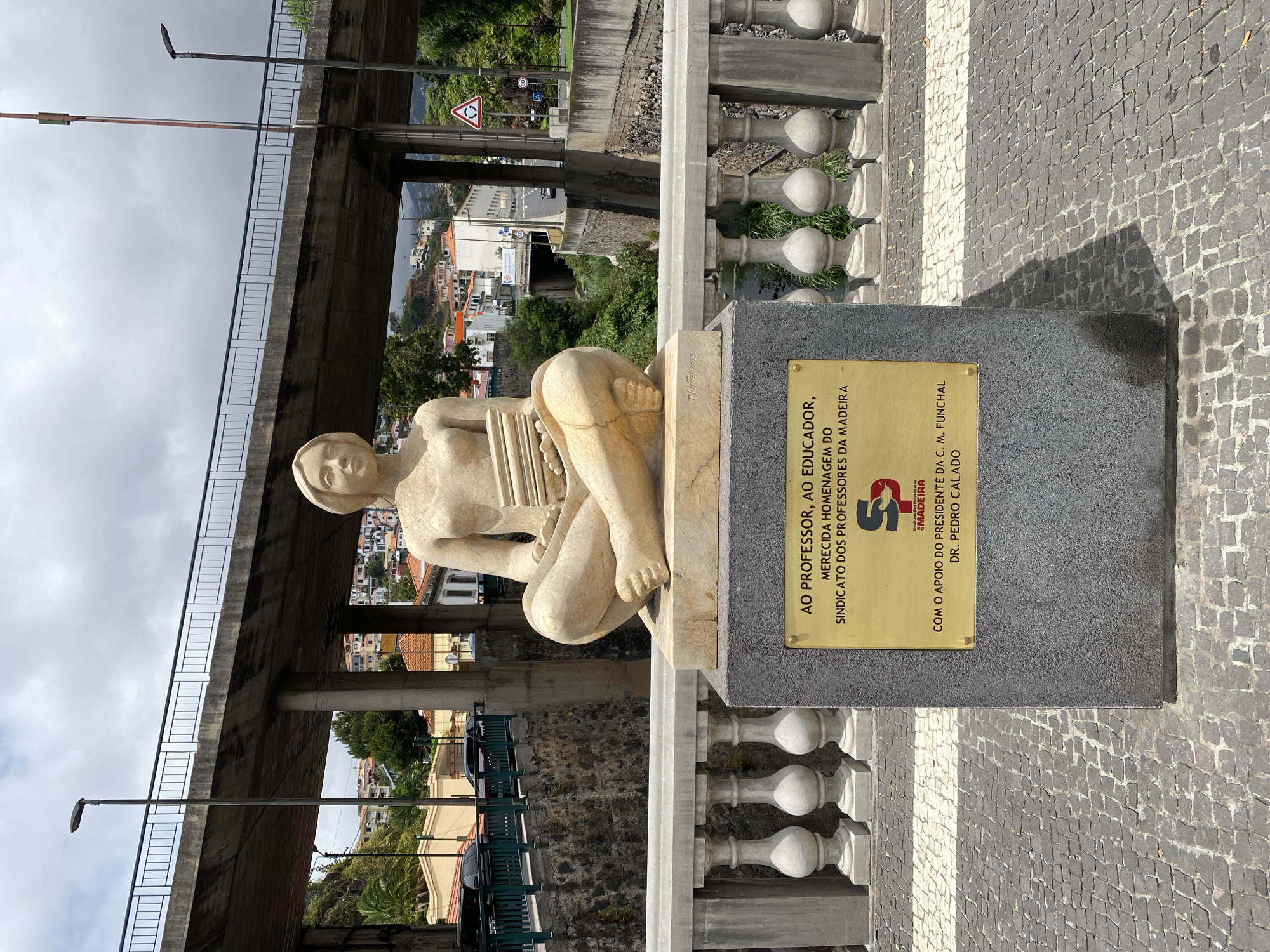 A stone sculpture of a seated figure holding books sits on a concrete pedestal on a bridge in Funchal, Portugal. The sculpture is a light beige color and depicts a person with a serene expression. The pedestal has a gold plaque with Portuguese text, indicating the sculpture is dedicated to professors and educators. Behind the sculpture, a concrete overpass spans across the scene. In the background, residential buildings with red roofs are visible on a hillside, characteristic of Funchal's urban landscape. The area around the pedestal is paved with cobblestones.