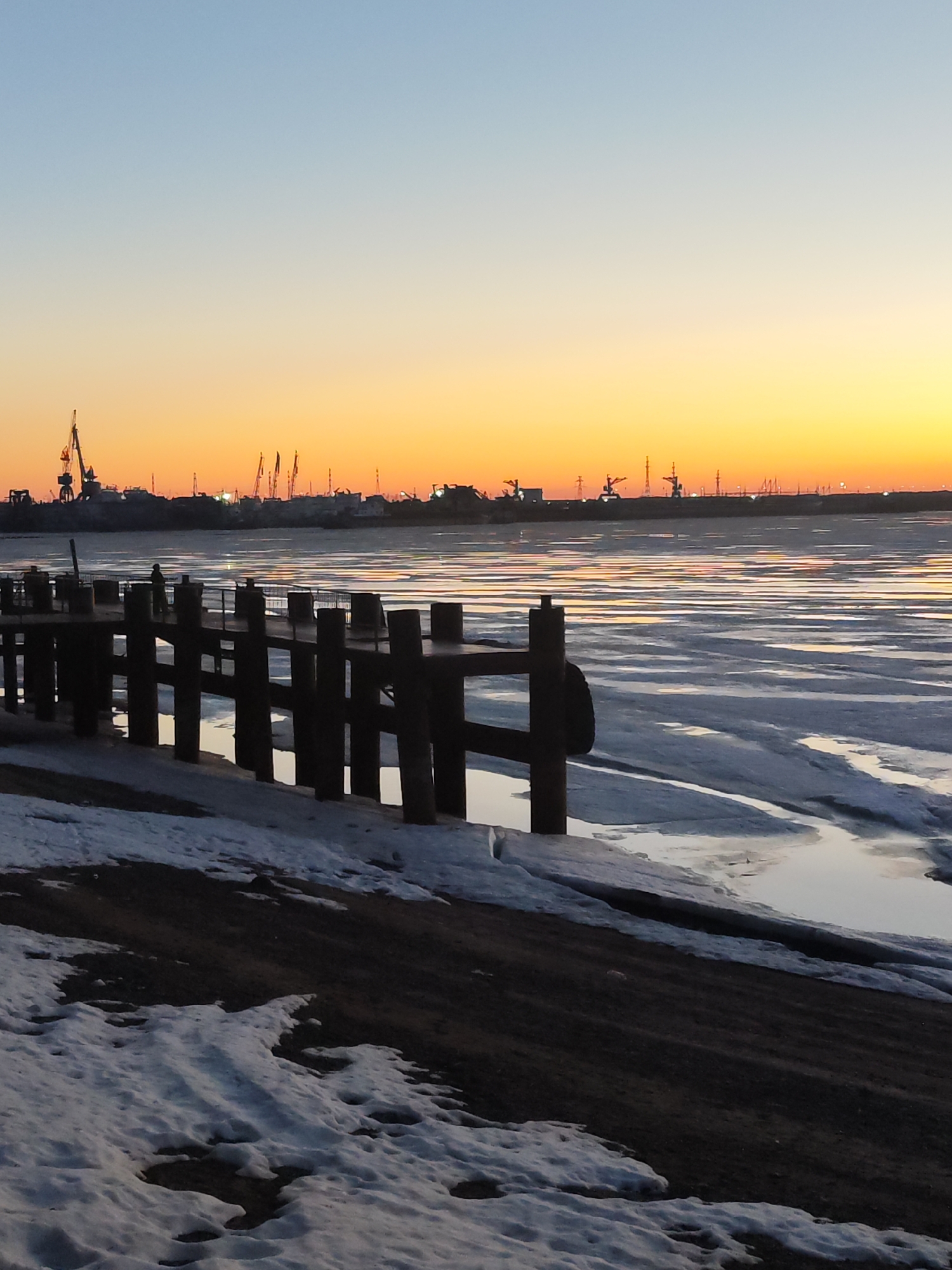 The image depicts a winter sunset over the Kama River in Chaikovsky, Russia. The foreground shows a partially snow-covered riverbank with a wooden pier extending into the icy water. The river is partially frozen with open water visible. In the background, the silhouette of industrial cranes and buildings of the industrial area is visible against the orange and yellow sunset sky. A lone person stands on the pier.
