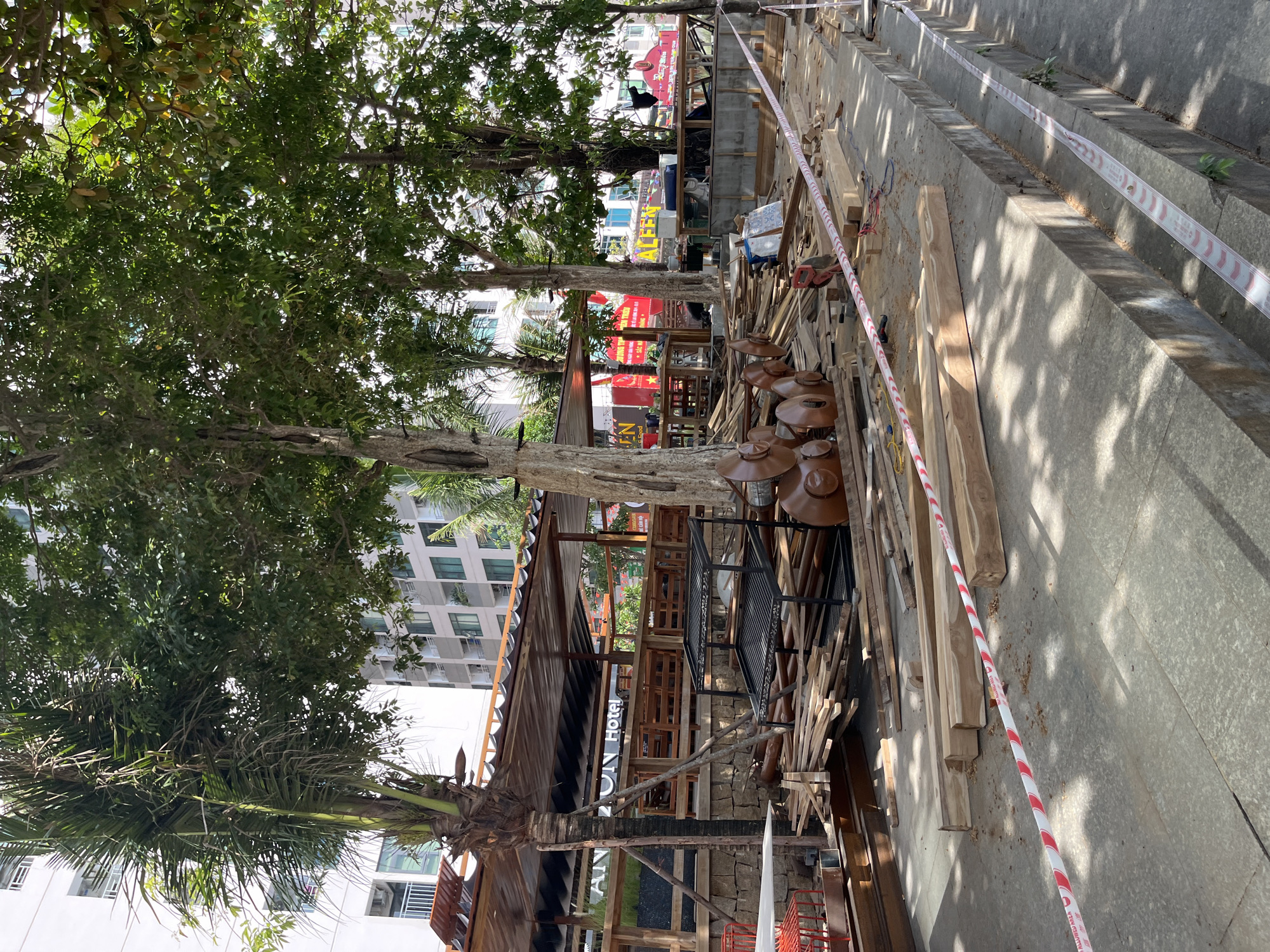 The image depicts a construction site with unfinished wooden structures and materials in the foreground. A partially constructed outdoor seating area or restaurant is visible in the background, with a sign that reads "AN NI Hotel". The scene is situated in an urban environment, as indicated by the presence of tall buildings behind the construction area. Red and white caution tape is laid out across the ground, suggesting a restricted or hazardous zone. The overall impression is one of ongoing construction or renovation work in a public area.