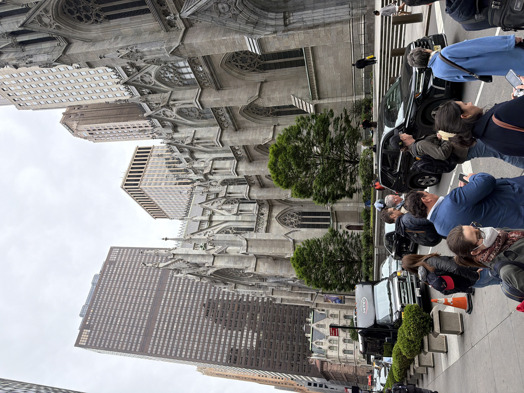 The image shows a street view in New York City, featuring St. Patrick's Cathedral, identifiable by its Gothic Revival architecture and towering presence. Adjacent to the cathedral are modern skyscrapers, creating a juxtaposition of architectural styles. A line of vehicles is parked along the side of the street, with pedestrians standing and observing the cathedral. Some people appear to be taking photos or videos, suggesting they are tourists or admirers of the architecture. There is foliage and urban landscaping visible, including trees and shrubbery. The overall scene is a mix of historical and contemporary elements typical of Manhattan's urban landscape.