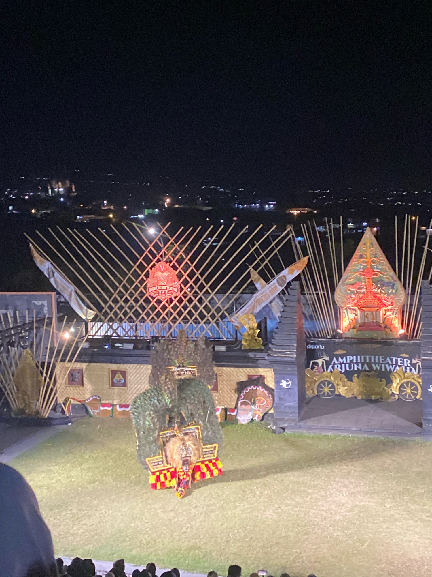An outdoor amphitheater stage is illuminated at night, set against a backdrop of a distant city skyline dotted with lights. The stage features elaborate decorations with a traditional Indonesian aesthetic. A large, stylized headdress with greenery and what appears to be animal fur or a mask is centrally positioned on the stage. To the right, a tall, pointed, red and gold structure, reminiscent of a temple or gateway, is lit from within. A banner above the stage entrance reads "AMPHITHEATER ARJUNA WIWAHA". In the foreground, an audience is partially visible, silhouetted against the stage lights. The ground in front of the stage is a grassy area, with shadows cast by the stage structures.