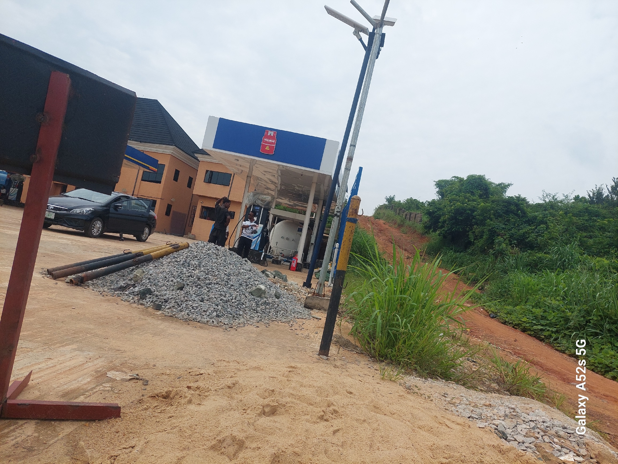 The image depicts a construction site and a fuel station in Ogbunike, Nigeria. In the foreground, a pile of gravel is visible next to a sandy area. Several large pipes, likely part of the construction, are laid out on the ground. Further back, a fuel station with a distinctive blue and white structure stands, featuring the logo of "OANDO". A black car is parked near the station, and a few individuals are present, some appearing to be working. The background shows buildings and lush greenery, indicative of the tropical environment. The sky is overcast. The image also displays a watermark in the lower-left corner: "Galaxy A52s 5G".