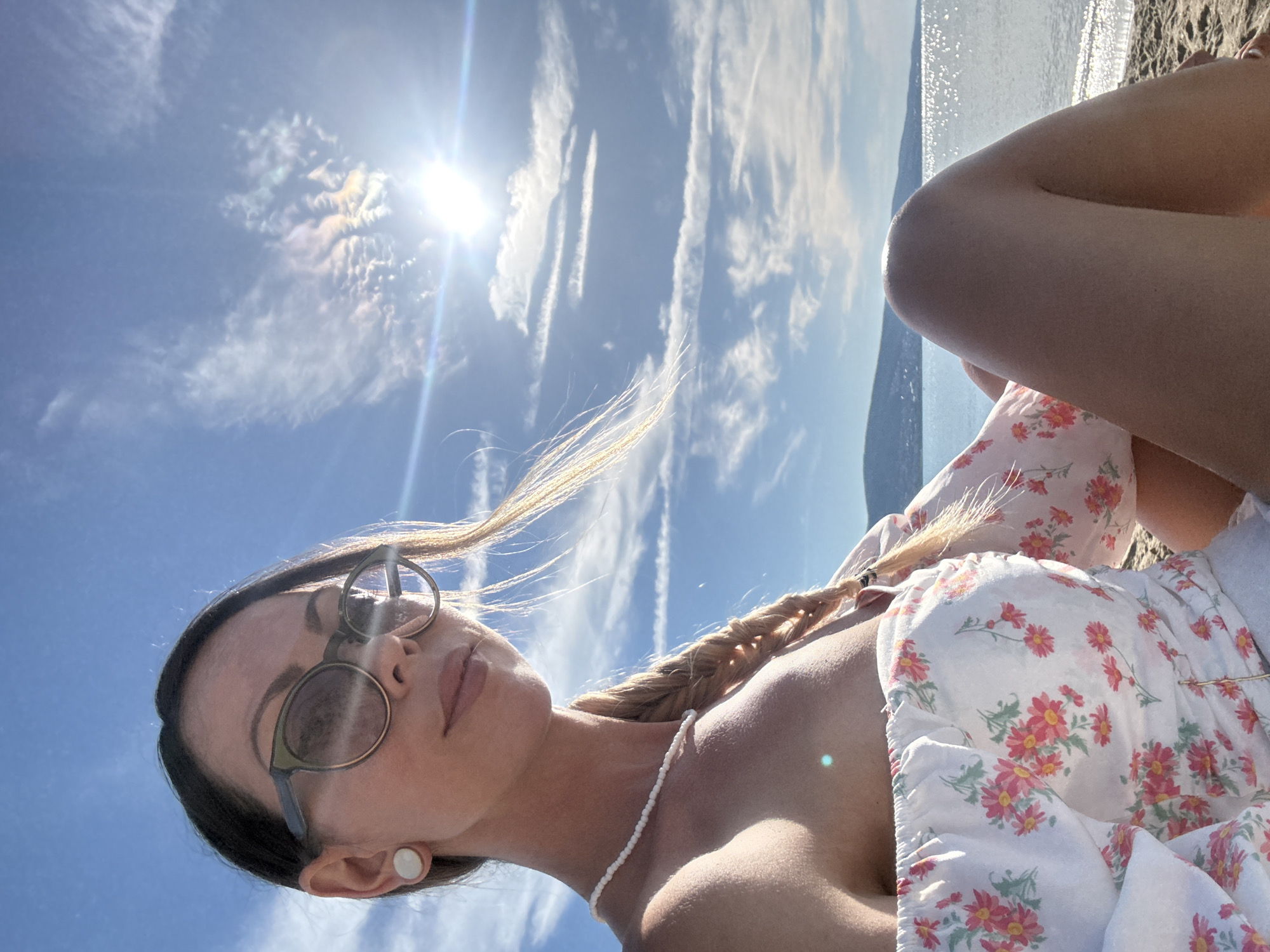 A woman is sitting on a beach in Nesebar, Bulgaria. She is wearing a white dress with a floral pattern, sunglasses, and a pearl necklace. Her hair is in a braid. The sun is shining brightly, and the sky is blue with some clouds. The sea is visible in the background.