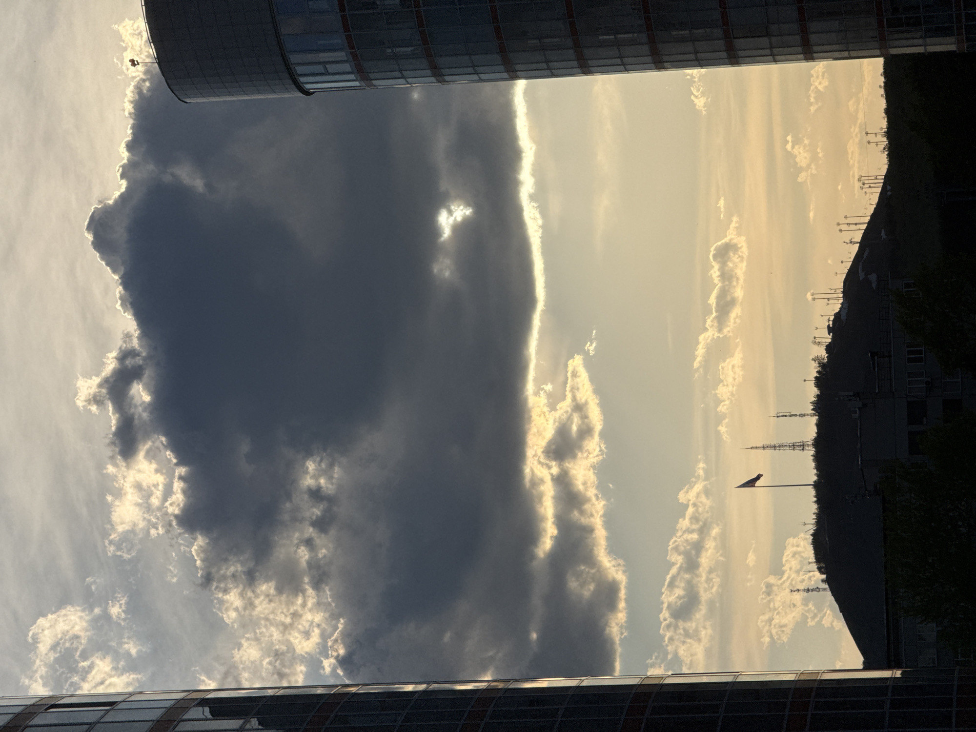 The image, oriented vertically, is framed by two modern buildings with glass facades at the top and bottom. The central view captures the sky dominated by a large, dark cloud formation to the left, juxtaposed against a brighter, sunlit area filled with cumulus clouds to the right. On the right, a silhouette of a hill with antennas is visible, as well as a flag. This panorama is likely a view over the city of Krasnoyarsk, Russia.