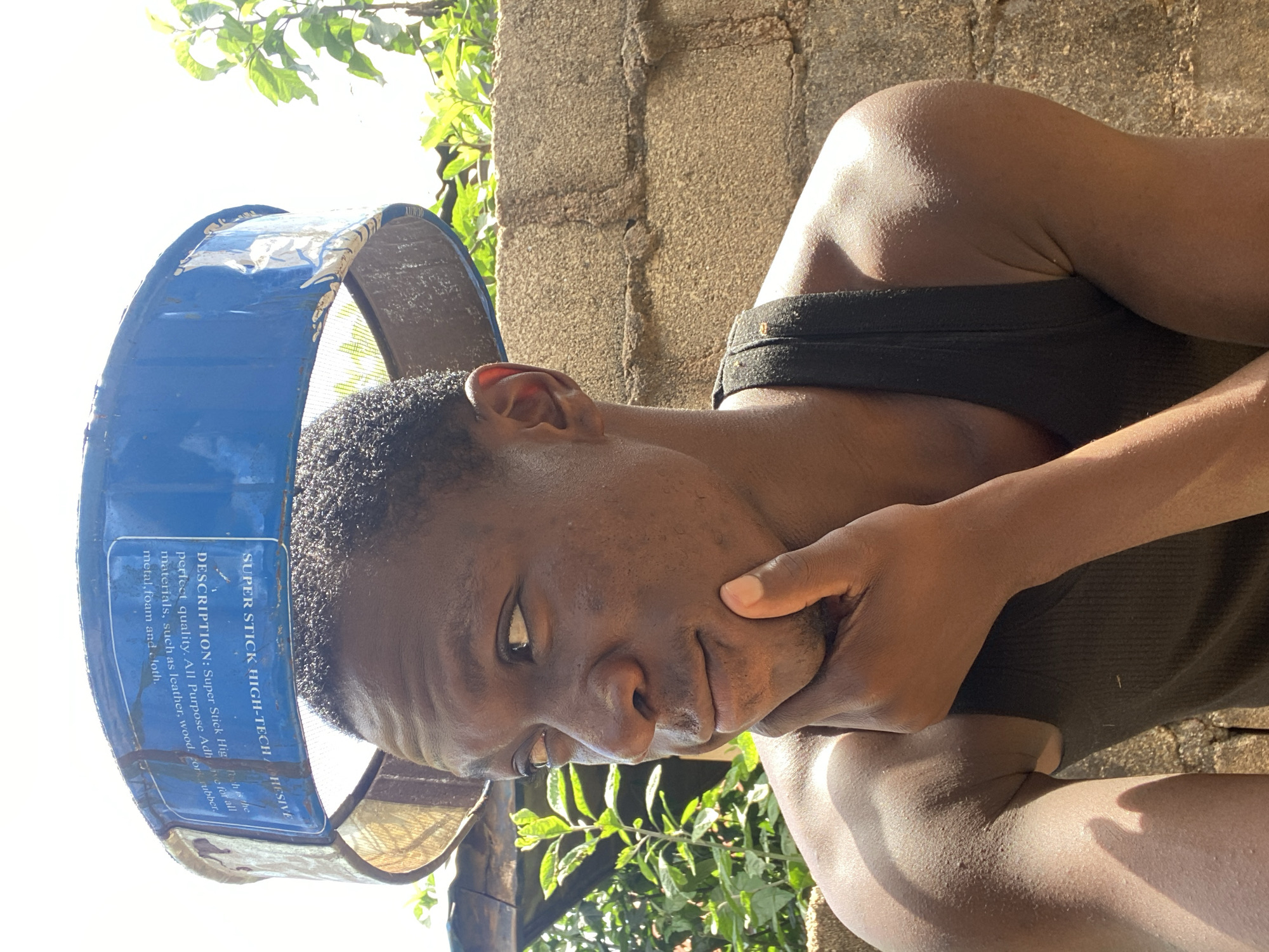 A young Black male is photographed outdoors in Bukuru, Nigeria, with his chin resting on his hand, looking to his left. He is wearing a black tank top and has short, dark hair. A blue metal rim with white lettering is positioned above his head, appearing to be part of a sieve or a similar tool. The background consists of a rough, textured brick wall and lush green foliage, with a bright, overcast sky above. The lighting suggests it is daytime. The overall scene conveys a candid, everyday moment.