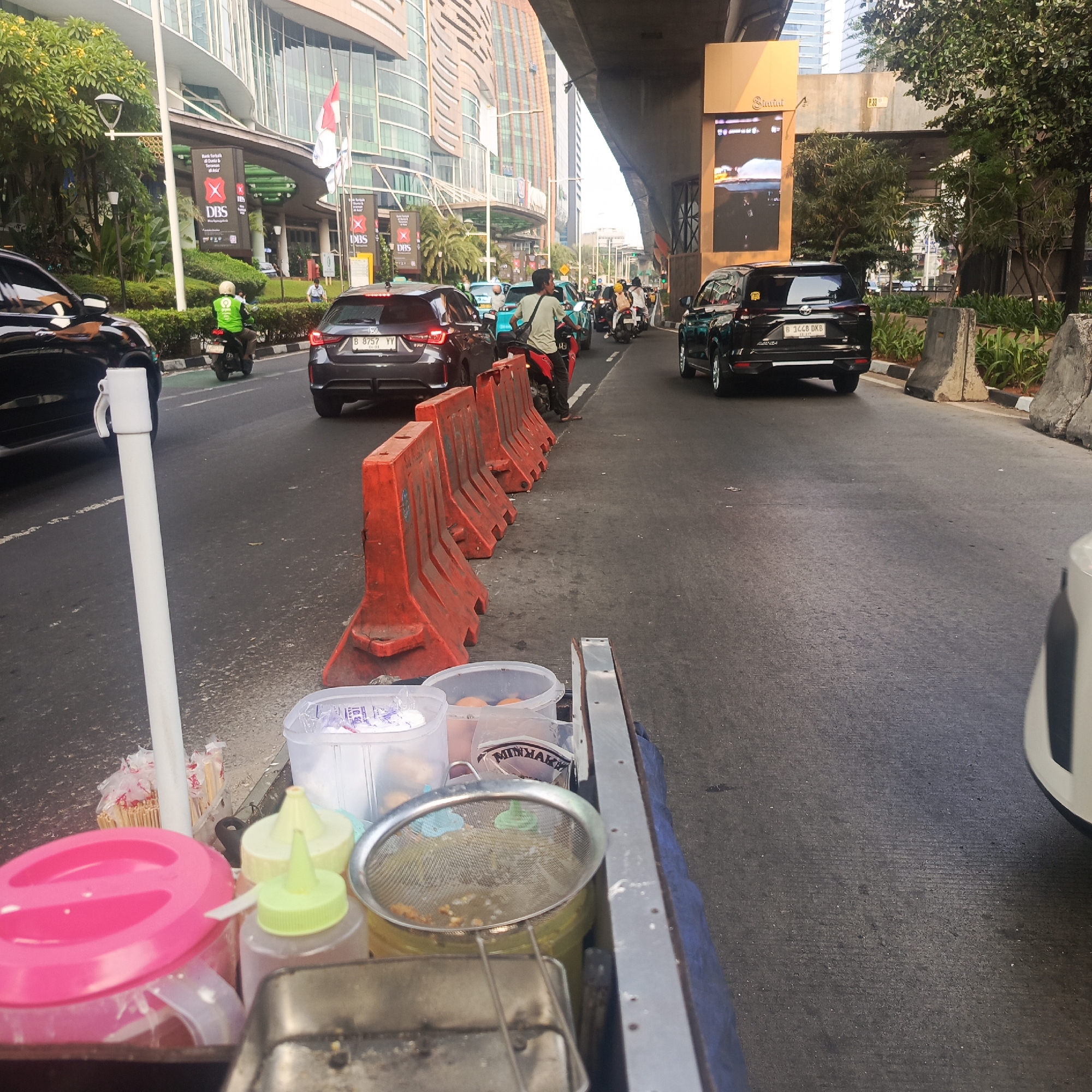 The image captures a street scene in South Jakarta, Indonesia, likely during daylight hours given the bright ambient light and clear visibility. The perspective is from behind a food cart, looking towards traffic on a multi-lane road.
In the foreground, the food cart is partially visible, with various items including a large pink container, bottles of condiments with yellow and green caps, a metal sieve containing food scraps, and other containers with food items such as eggs and what appears to be a package of seasonings. Skewers are also visible in a cylindrical container. A white pole, possibly part of a temporary barrier or signpost, stands to the left of the cart.
The road itself shows moderate traffic congestion. Several vehicles are present, including a black SUV with license plate "B 1448 DKB", a dark-colored hatchback with license plate "B 8757 YY", a black car on the far left, and other cars and motorcycles further down the road. One motorcycle rider, wearing a bright green jacket and white helmet, is seen in the middle lane. The road is flanked by orange traffic barriers on the right side and concrete barriers and greenery on the far right.
In the background, tall modern buildings with glass facades dominate the skyline, indicative of a commercial or business district. A large digital billboard is visible on the right side of the image, displaying an advertisement. An elevated road or bridge structure passes over the scene. Indonesian flags are visible on poles in front of one of the buildings. The overall impression is a busy urban environment with ongoing commercial activity and traffic flow.