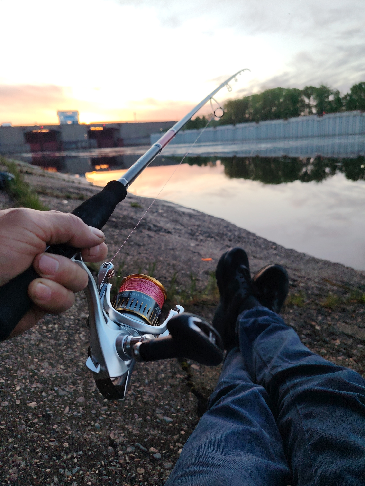 The image shows a person fishing on a riverbank at sunset in Chaikovsky, Russia. The person is wearing dark shoes and blue pants and is holding a fishing rod with a silver reel with red line. The fishing line extends out into the water. The river is calm, reflecting the sunset colors. In the background is a concrete structure, possibly a dam or retaining wall, with green trees beyond. The ground is rough concrete. The reel is labeled "Fiber Pro 3000".