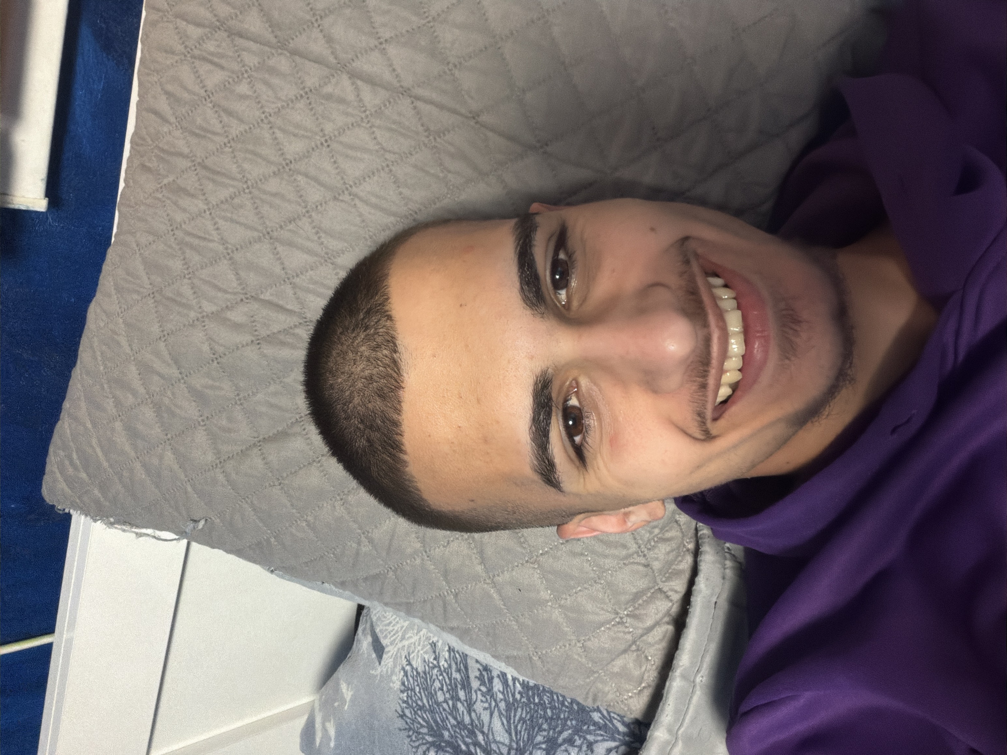 A man with a shaved head, dark eyebrows, and brown eyes is smiling into the camera. He is wearing a purple hooded sweatshirt and is lying on a gray quilted fabric. A portion of a white object and a blue/gray patterned fabric are also visible in the background. No information can be gathered about the location in Sofia, Bulgaria.