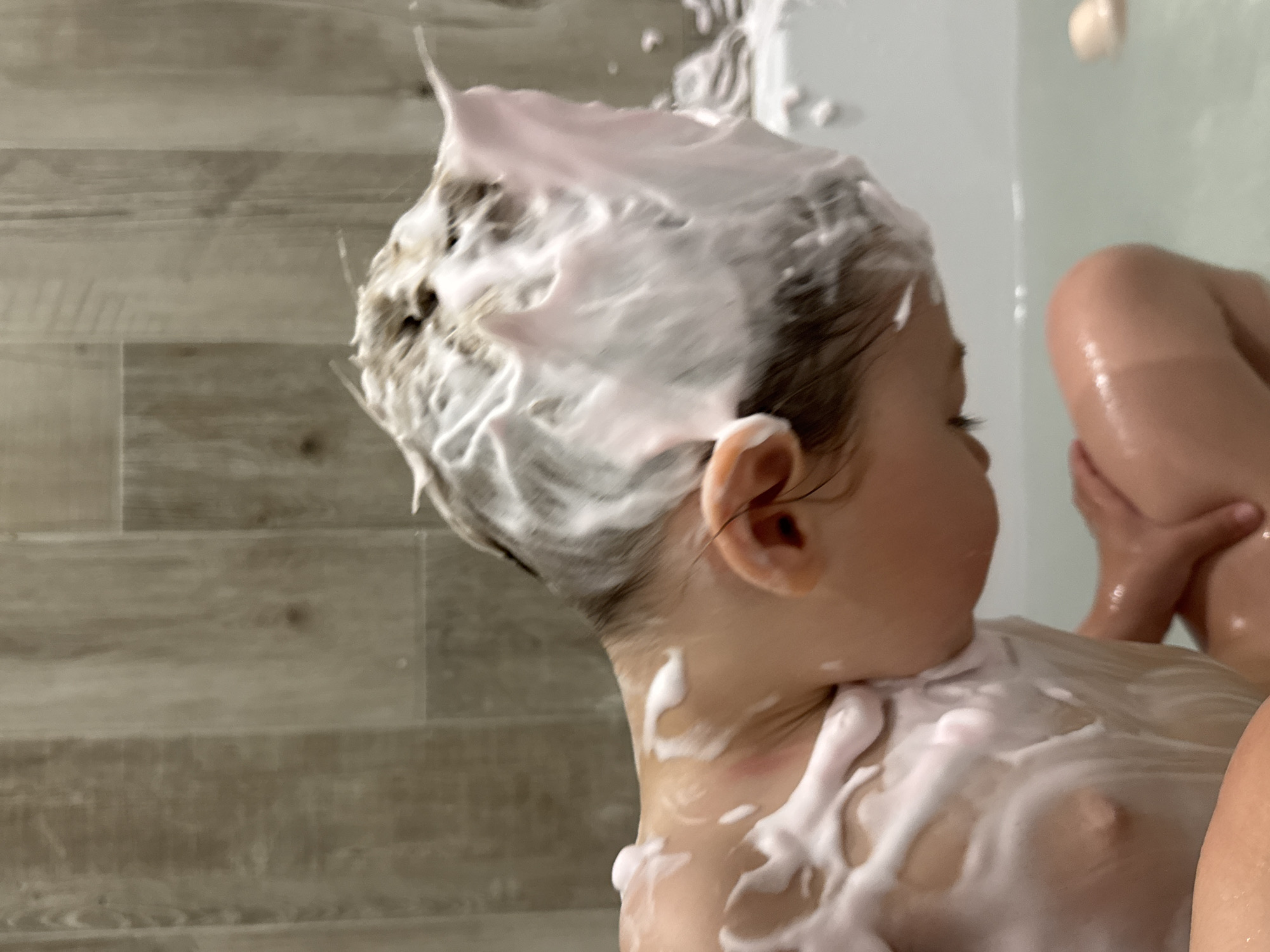 A close-up, slightly blurred photograph shows a child in a bathtub covered in pink and white foam. The child is facing away from the viewer, with their head turned slightly to the right. Foam covers the child's head, face, and chest. The child's ear is visible. In the background, there's a blurry image of a wall with wood-like paneling. Part of another person's body, possibly an arm and leg, is visible in the upper right of the frame. The photo was possibly taken in Zelenogradsk, Russia.