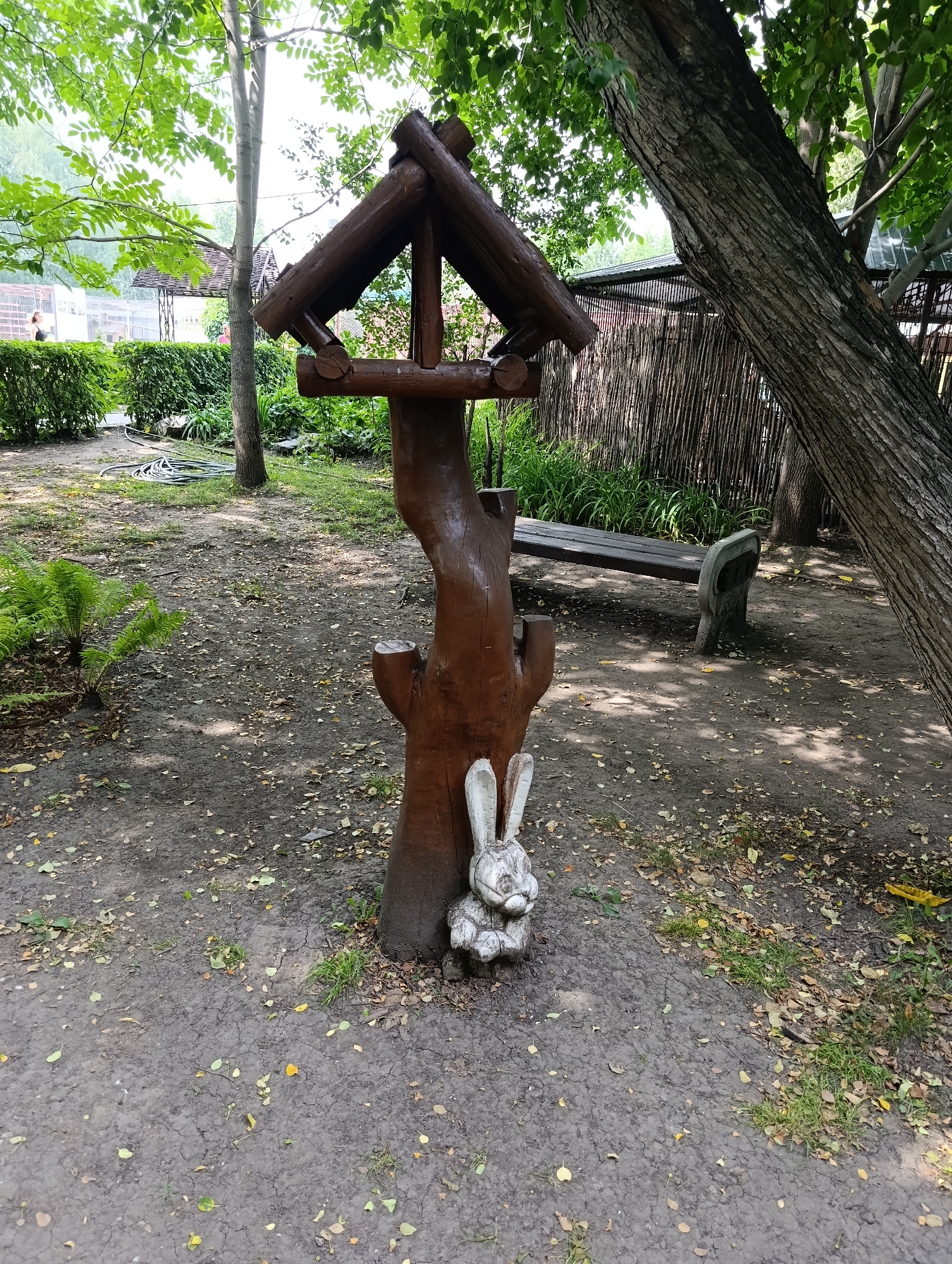 The image depicts an outdoor scene, possibly in a park or garden setting. A rustic wooden bird feeder, constructed from logs, stands prominently in the center. Adjacent to the base of the bird feeder is a small, weathered statue of a rabbit. In the background, a wooden bench is visible, and beyond that, a fence made of vertical sticks. Trees and greenery surround the area, with fallen leaves scattered on the ground. The lighting suggests it is daytime, with shadows cast by the trees. There are no people clearly visible in the immediate vicinity of the bird feeder and rabbit statue. Without additional context or identifiable landmarks, it is not possible to determine the specific location within the city of Barnaul, Russia.