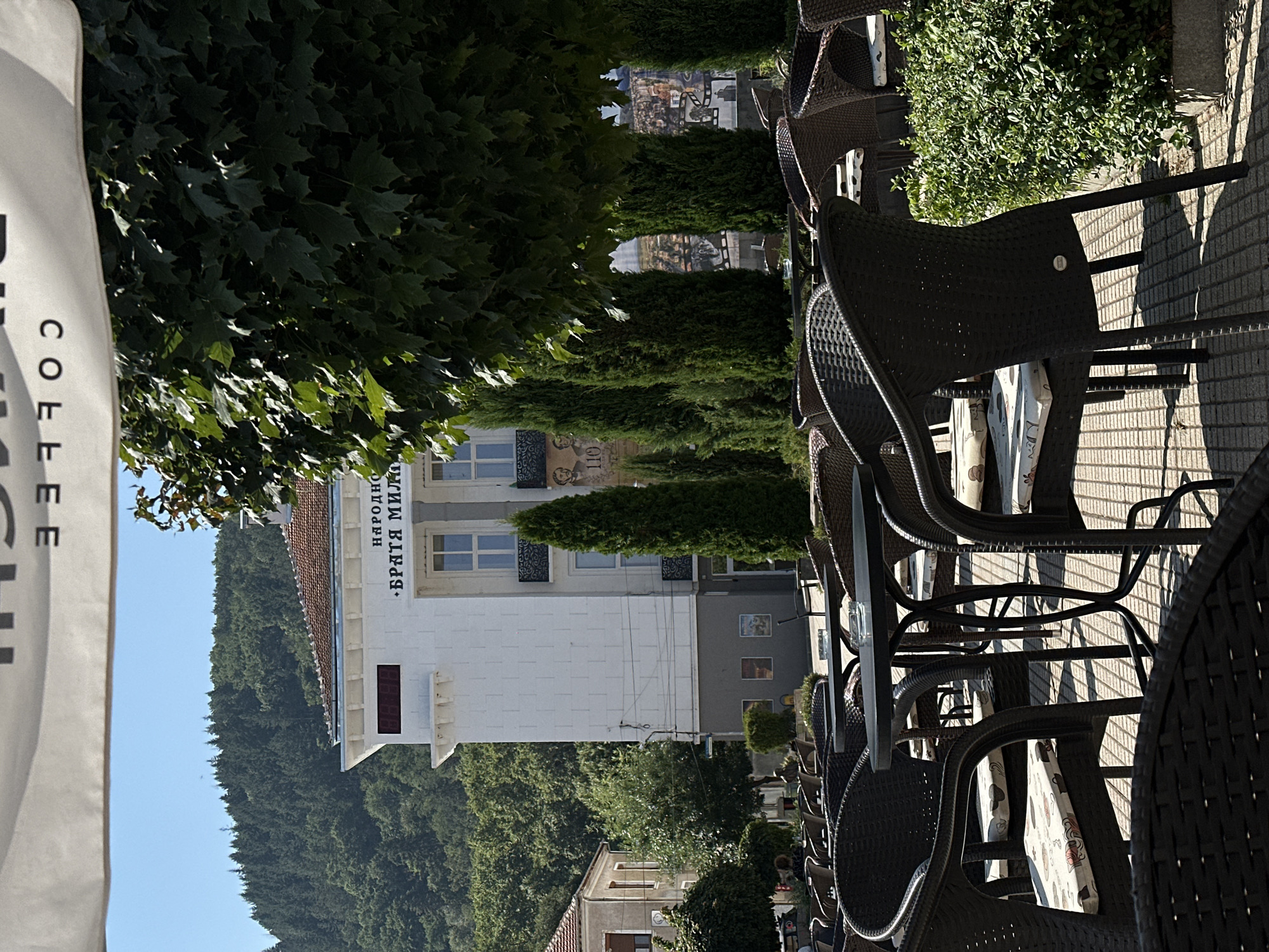 An outdoor cafe in Petrich, Bulgaria, on a sunny day. The foreground features several dark wicker chairs and tables set up on a tiled patio. Cushions with a patterned design are placed on the seats of the chairs. In the middle ground, a white building with a red-tiled roof is visible, bearing Cyrillic text that reads "НАРОДНО ... БРАТЯ МИЛАНОВИ" (National ... Brothers Milanovi). A digital clock displaying "88:88" is mounted on the facade of the building. Several tall, conical evergreen trees are positioned in front of the building. The background shows a densely forested hill under a clear blue sky.