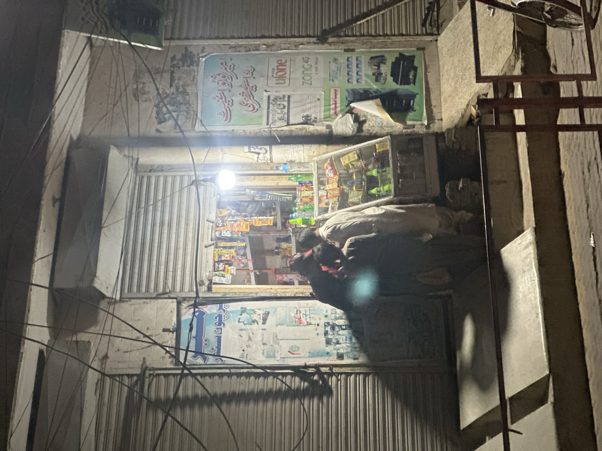 The image depicts a nocturnal scene outside a brightly lit, small shop or general store. The shop is set within a building with concrete walls, flanked by closed metal roll-down shutters of neighboring establishments. Numerous dark electrical wires are visible, hanging across the frame.
Inside the shop's open entrance, shelves are densely packed with merchandise, including various snacks, drinks in plastic bottles, and other general goods. A single, prominent light fixture inside the shop illuminates its interior and casts a glow onto the area immediately in front of it.
Two individuals are present at the shop's entrance. One person, wearing a light-colored garment, is standing facing inwards, looking at the items within the shop. The second individual, dressed in darker clothing, is hunched over with their back partially to the camera, seemingly engaged in an interaction or transaction with the shop or the other person.
Above the shop's opening, a large sign is displayed. Visible text on this sign, presented in Urdu and English, includes "امیر فوٹو اسٹیٹ" (Amir Photo State), "پرچون" (Retail/General Store), "یو فون" (Ufone), "زونگ 4G" (Zong 4G), "بیٹا فلیکسی" (Beta Flexi), and "ADAA". Below the main entrance, a faded poster with indistinct text is also visible. The scene suggests a typical evening transaction at a local shop in an urban setting.