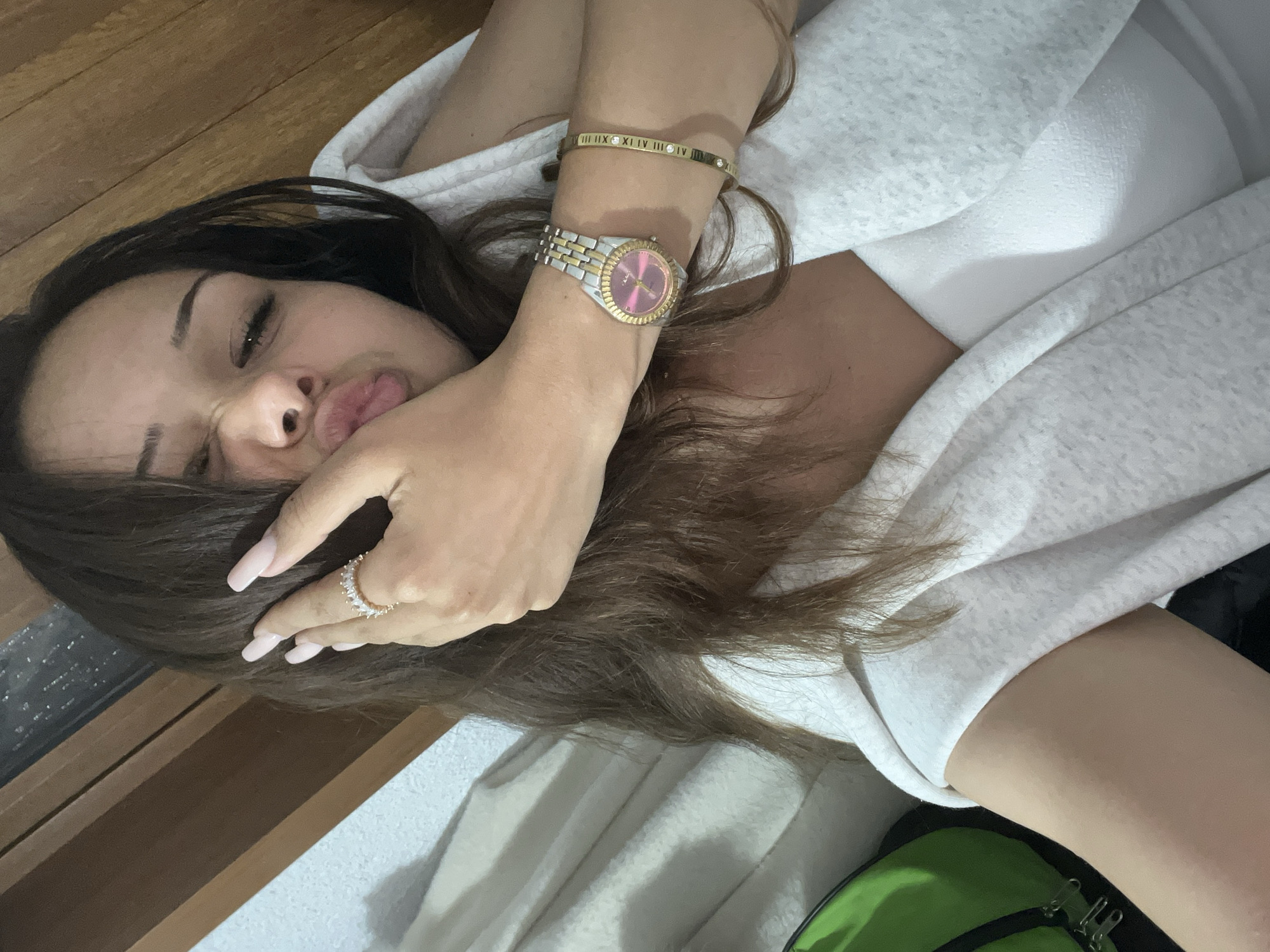 A young adult female is depicted in a close-up, upward-angle selfie within an indoor private setting in Unknown, Unknown. Her long, wavy dark brown hair frames her face, partially obscured by her right hand, which displays long, light pink/nude manicured nails and a gold-toned ring with multiple clear baguette-cut stones on her ring finger. She is making a puckered lip expression with squinted eyes. She wears a light gray or white textured outer garment over a white inner top. On her left wrist, she wears a two-tone (silver and gold-toned) watch with a pink dial featuring gold-toned Roman numeral hour markers, and a thin gold-toned bangle bracelet with engraved Roman numerals and interspersed clear stones. The background on the right consists of vertical wooden planks, while the left features a lighter textured wall or fabric. A portion of a green backpack with silver zippers is visible in the lower left corner.