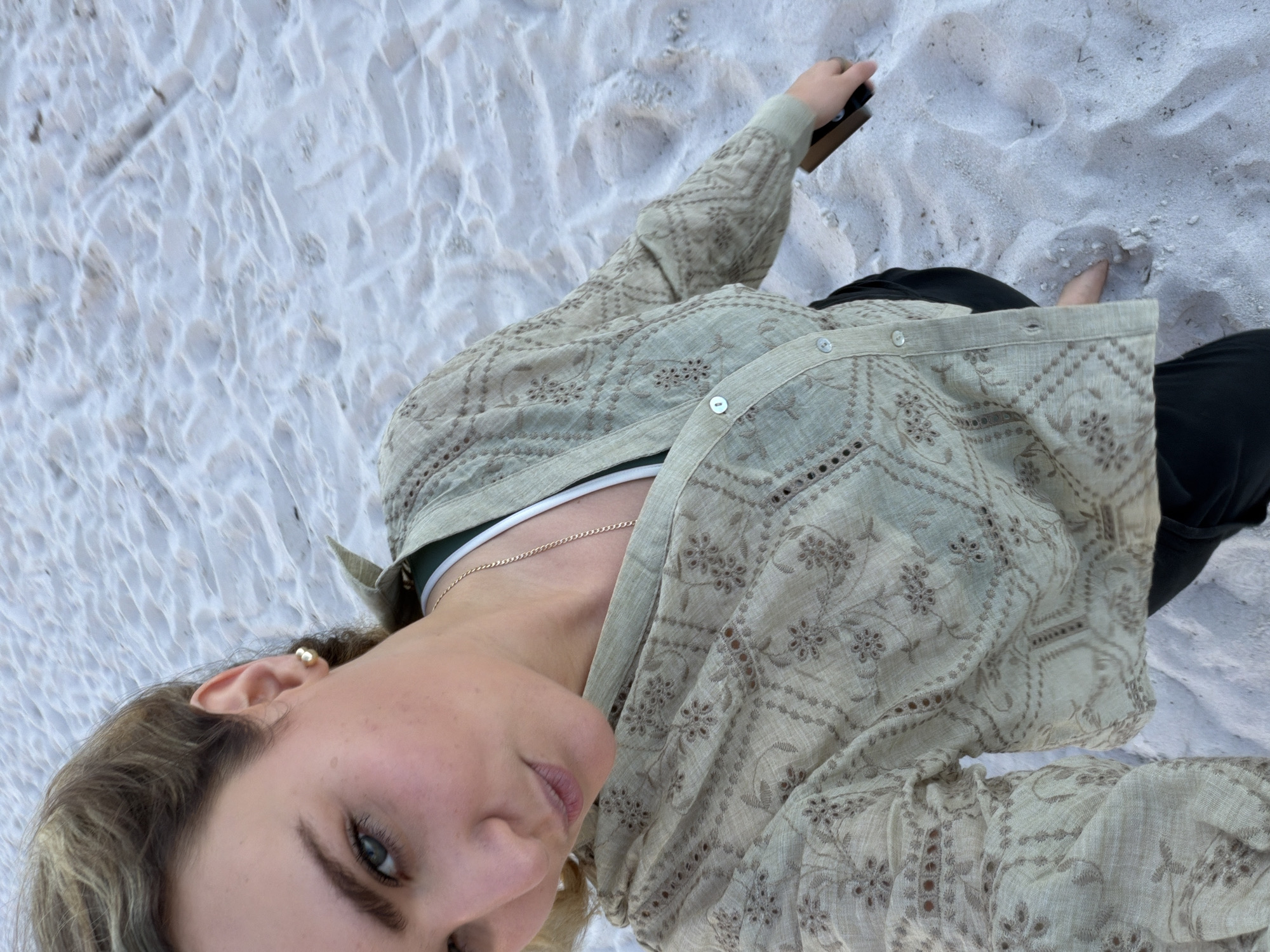 A person is lying on a beach in Key Biscayne, Florida, United States. The person has blonde hair and is wearing a light green patterned shirt with black pants. The person is lying on white sand. The person is holding a phone in their right hand.