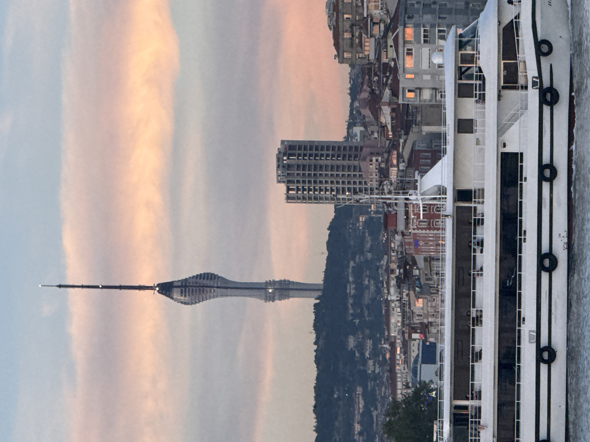 The image shows a view of the Çamlıca Tower and a modern multi-story building in Kadıköy, Turkey, set against a sky with wispy pink and orange clouds. In the foreground, a white, multi-deck ferry or cruise ship is moving across the water, with several people visible on its decks. The ship has a red waterline and black tires mounted along its sides. Behind the ship, a dense urban landscape of residential buildings with red-tiled roofs and varying architectural styles fills the midground. The Çamlıca Tower, a prominent landmark, is visible in the distance, towering over a tree-covered hill. The overall scene depicts a coastal urban environment during either sunrise or sunset.