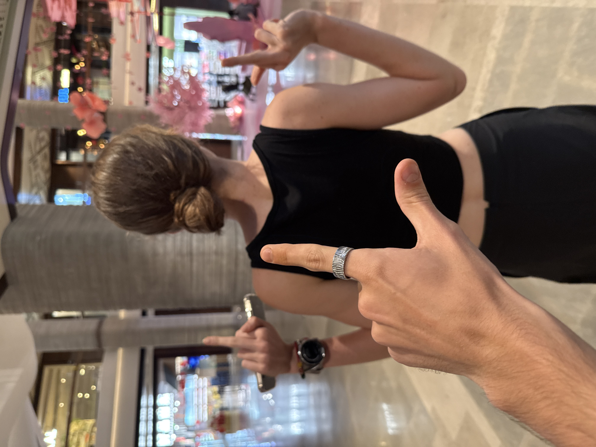The image, originally rotated 90 degrees counter-clockwise, depicts an indoor scene likely within a commercial complex or shopping mall in Putuo District, China.
In the immediate foreground, the right arm and hand of one person are prominently featured. Their index finger points directly towards another individual, and a distinctive silver-toned ring with a textured, wavy pattern adorns this finger. The foreground person's lower chest and midriff, clad in a black top, are also partially visible.
In the mid-ground, a second individual is seen from behind and slightly to the side, with their head turned away from the camera. This person has light brown hair styled in a neat bun and is wearing a black sleeveless or crop top and dark bottoms. They are actively making a dynamic hand gesture with both hands, fingers splayed outwards. A watch is visible on their left wrist, and they hold a silver-colored smartphone or similar device in their left hand, also pointing with their index finger.
The background is intentionally blurred, creating a bokeh effect that highlights the subjects. Despite the blur, elements consistent with a modern, well-lit interior are visible. Pink, floral-like decorations are suspended or arranged throughout the space, possibly as part of a seasonal display or an artistic installation. Glimpses of illuminated storefronts or displays with various lights can be seen in the soft-focus distance, suggesting a bustling commercial environment. The floor is a light-colored, reflective surface, possibly tiled or polished concrete.
The activity appears to be a casual or playful interaction, possibly a posed photo op, characterized by the hand gestures and pointing. The lighting suggests a bright indoor setting, whether daytime or a well-illuminated evening. There is no discernible text visible in the image.