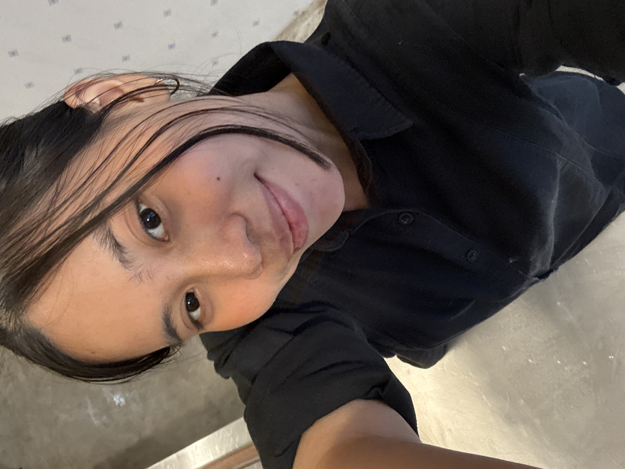 A young woman is taking a selfie indoors. She has dark hair and is wearing a black collared shirt. The background appears to be a wall with a subtle pattern and a light-colored surface below. The scene is likely in a residential or commercial space within South Jakarta, Indonesia, given the context of the image's origin.