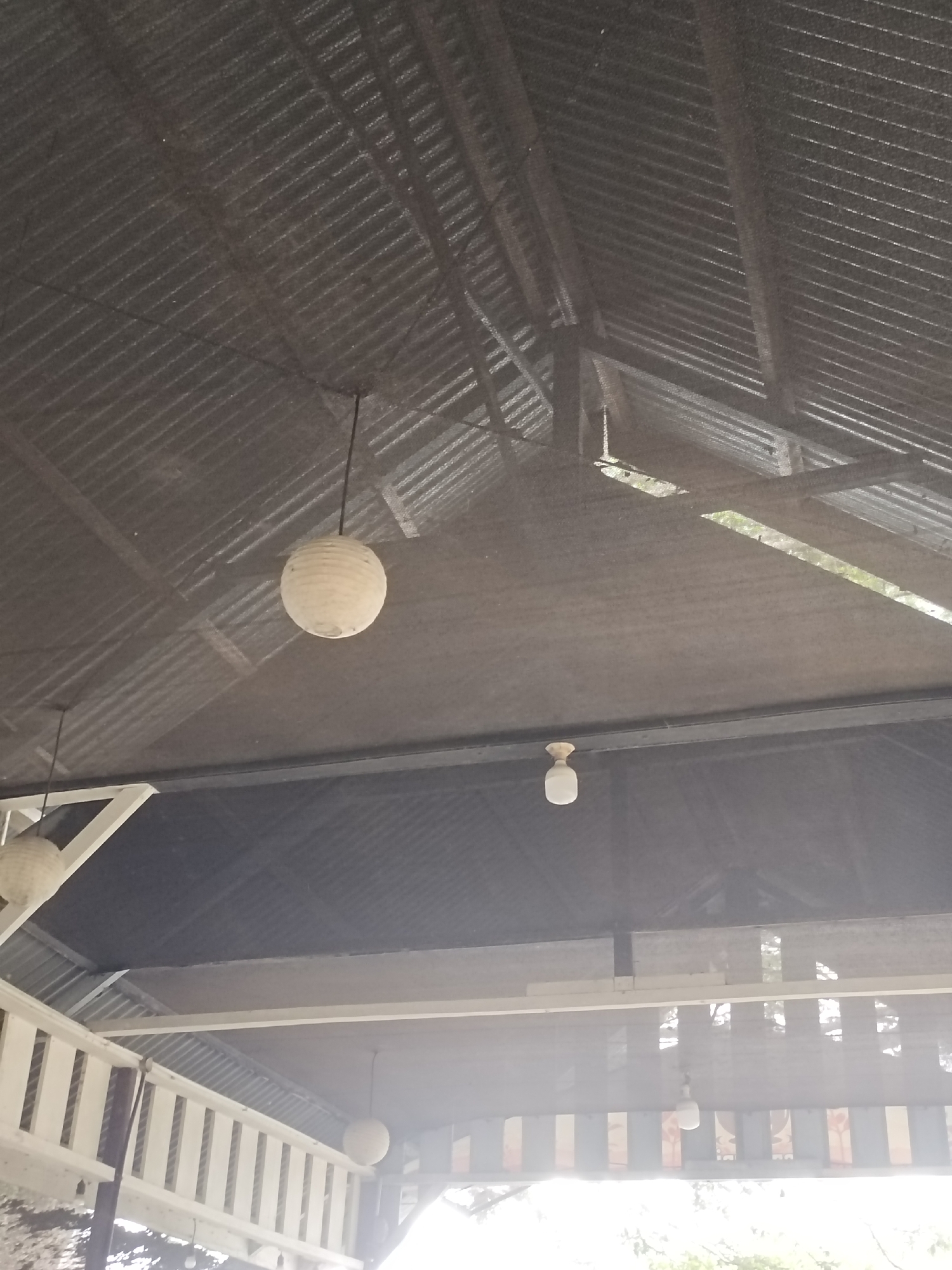 The image captures an upward view of a sheltered outdoor or semi-outdoor structure, likely a veranda, patio, or open-air restaurant, in Keudematangkuli, Indonesia. The dominant feature is the corrugated metal roof or ceiling, which appears dark grey and is supported by a network of visible wooden beams and trusses. A section of the ceiling is covered by a darker mesh or fabric material, possibly for shade or insect protection.
Several light fixtures are visible. In the upper left quadrant, a white spherical paper lantern hangs from the ceiling. A similar, partially visible lantern can be seen further to the left and lower down. To the right of the central lantern, a more conventional ceiling light fixture with a white bulb is attached to a beam. Another identical light fixture is visible lower in the frame, near the bottom right.
A white wooden railing or balustrade runs horizontally across the lower left portion of the image, contributing to the architectural structure of the space. The lower part of the image is brightly lit by natural sunlight, indicating it is daytime. Glimpses of green foliage, likely from trees, can be seen beyond the structure, particularly in the brightly lit areas, suggesting an outdoor setting with natural surroundings. There is no visible activity or people in the image, nor is there any discernible text. The scene conveys a sense of a functional, rustic, and shaded area.
