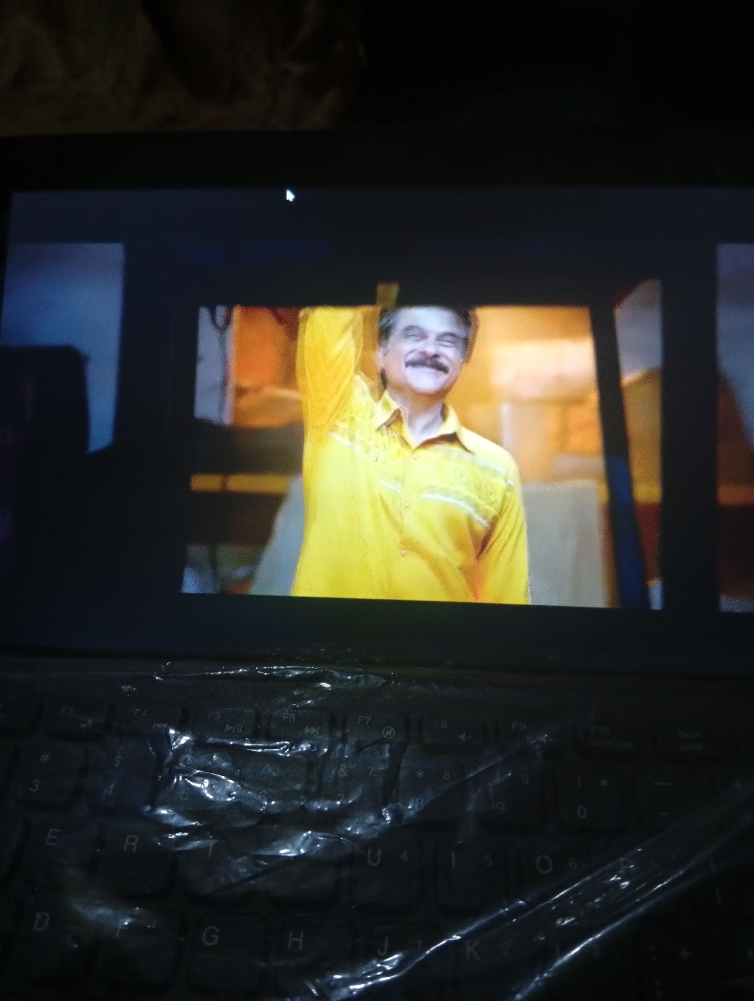 The image shows a man in a yellow shirt with a mustache, his right arm raised, smiling. He appears to be in an indoor setting with warm-toned walls and structures in the background. The image is taken from a slightly elevated angle, looking down at the man. The bottom portion of the image displays a black keyboard covered with plastic. The overall scene is dimly lit, suggesting it might be a photograph taken at night or in a low-light environment. The location is not identifiable from the image.