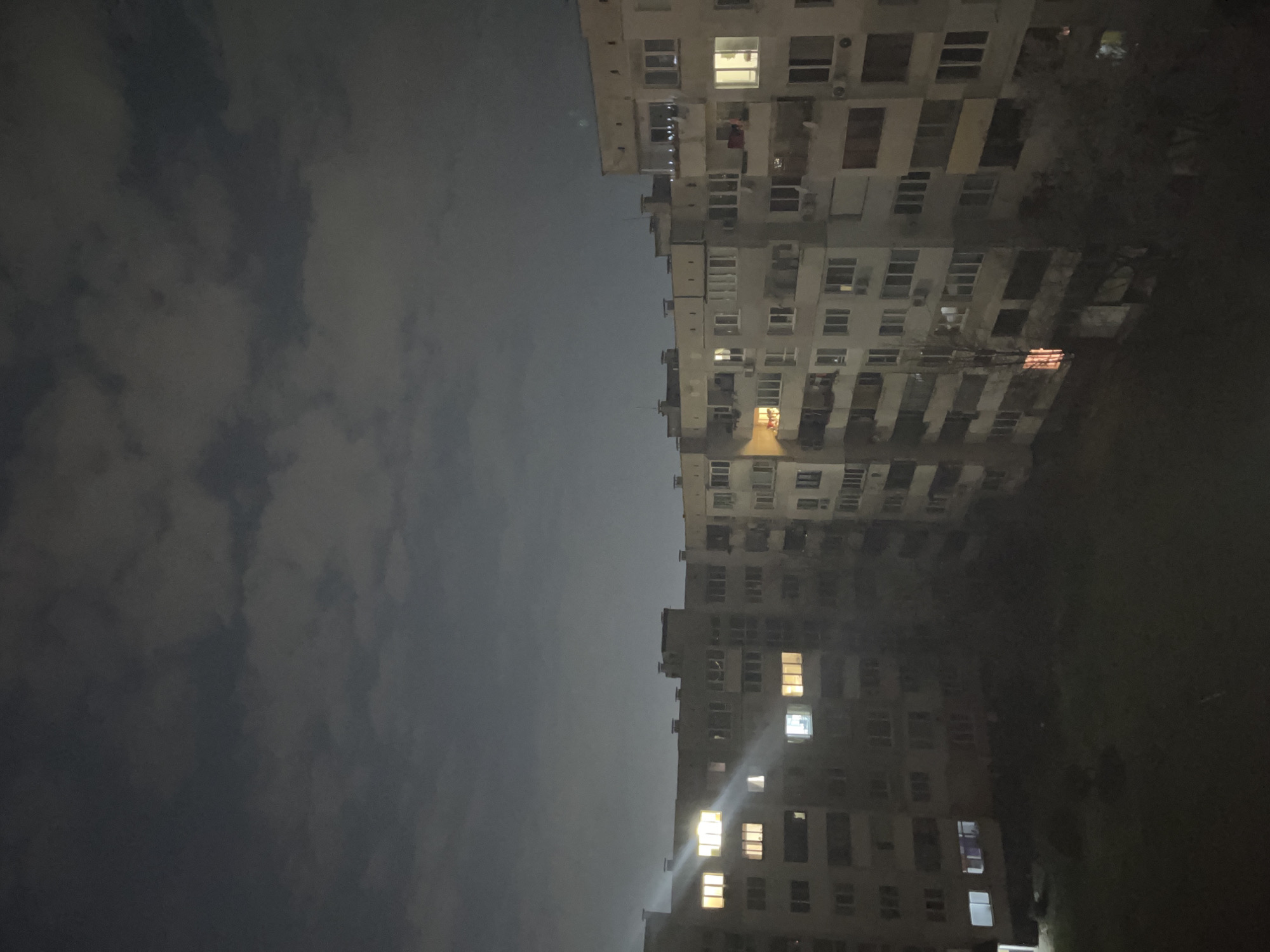 The image depicts a nighttime urban residential scene in Sofia, Bulgaria, characterized by multiple multi-story apartment buildings. The sky overhead is dark and mostly overcast, with indistinct clouds partially visible.
The foreground and midground are dominated by several beige-colored concrete apartment blocks, ranging approximately from seven to nine stories in height, arranged to form an inner courtyard or shared green space. The architecture is typical of socialist-era residential complexes common in Sofia. Numerous windows across the facades are illuminated with artificial light, indicating occupancy, while others remain dark. Some balconies are visible, with minor details such as clothes hanging or air conditioning units on exterior walls. A prominent bright light source emanates from an upper-floor window on the leftmost building, causing a noticeable lens flare extending upwards and to the right. The ground level directly below the buildings is dark and unlit, appearing as a sparsely vegetated area with some discernible trees or bushes. No people, vehicles, or specific street furniture are visible within the frame. The overall impression is one of quiet, late-night domesticity within a densely populated urban residential block.