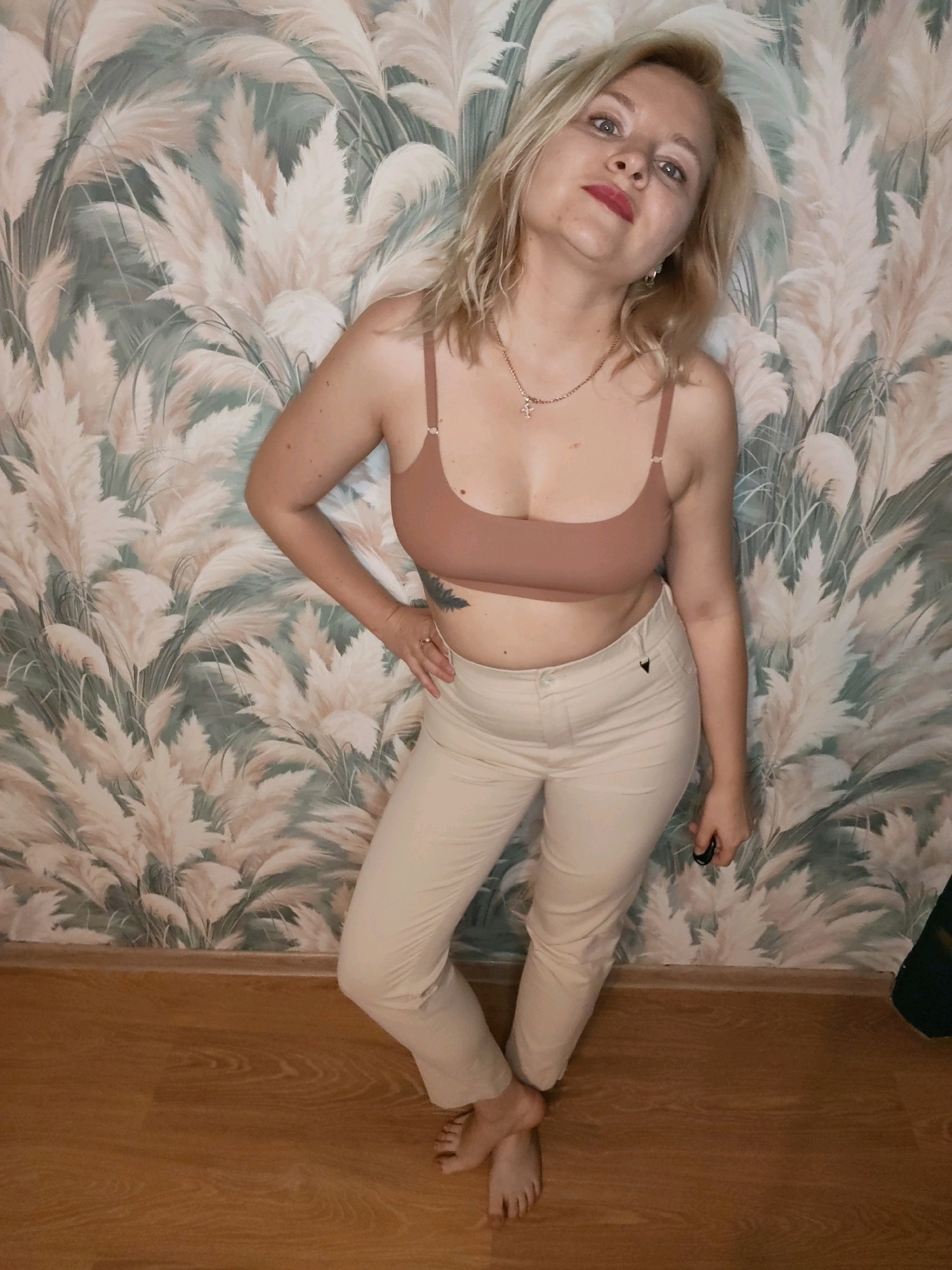 A full-body shot shows a fair-skinned woman with blonde hair and red lipstick, leaning against a wall with a pampas grass-patterned wallpaper. She is wearing a nude-colored bra and light beige pants. Her right hand is on her hip, and her left hand is holding a small black object. She has a tattoo on her left hip. The floor is made of light brown wood. The image appears to have been taken indoors during the day. The woman's expression is one of mild curiosity or amusement. There is no visible text in the image. The location context is Izhevsk, Russia.