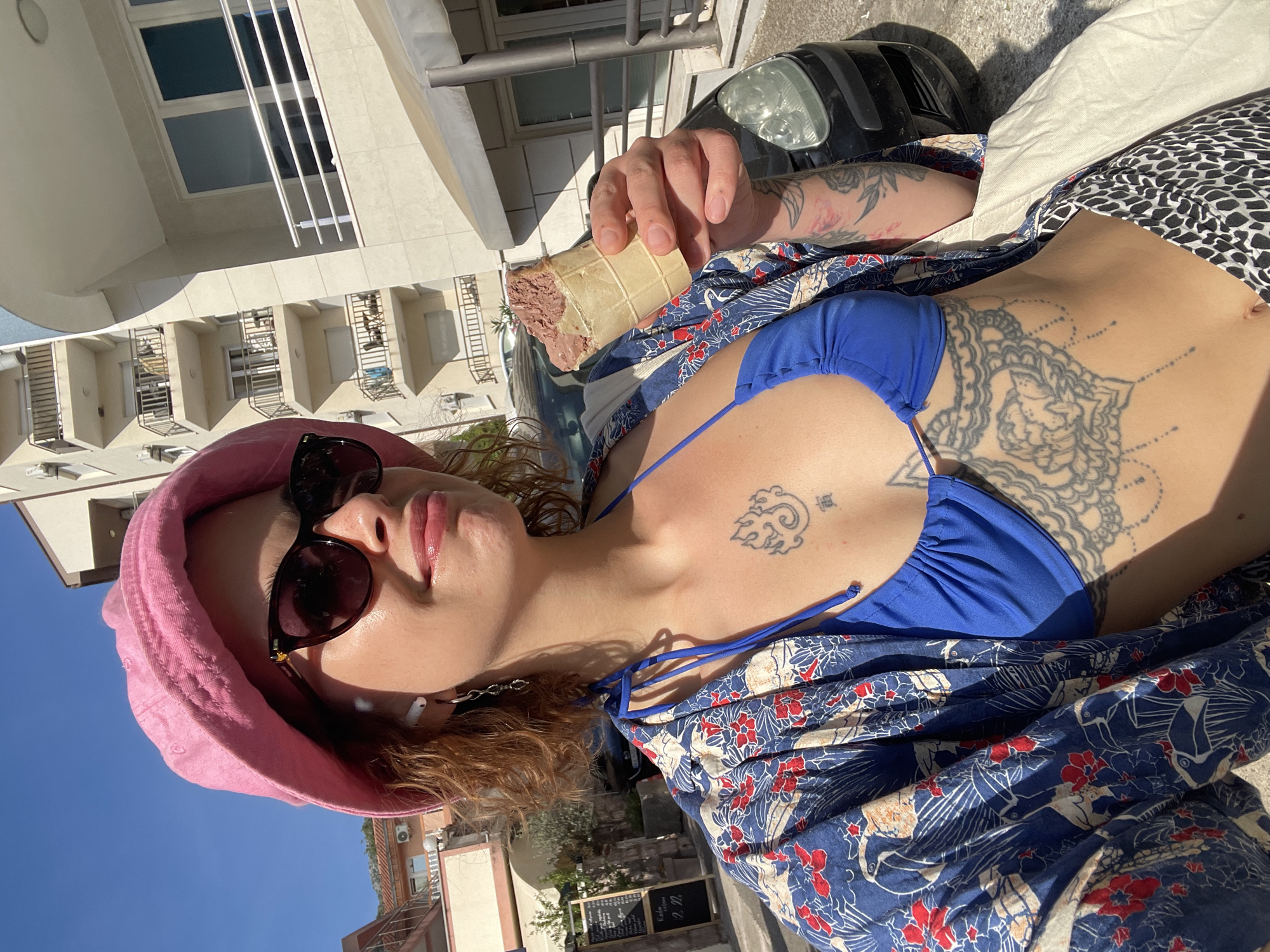 The image depicts a person lying down outdoors on a concrete surface, wearing a blue bikini top, a blue and white patterned shirt, a pink hat, and sunglasses. The person holds an ice cream cone. Tattoos are visible on their chest and arm. In the background, there is a multi-story building with balconies, a parked vehicle, and a menu board in front of a building. The location is Rafailovići, Montenegro, based on the architecture and signs.