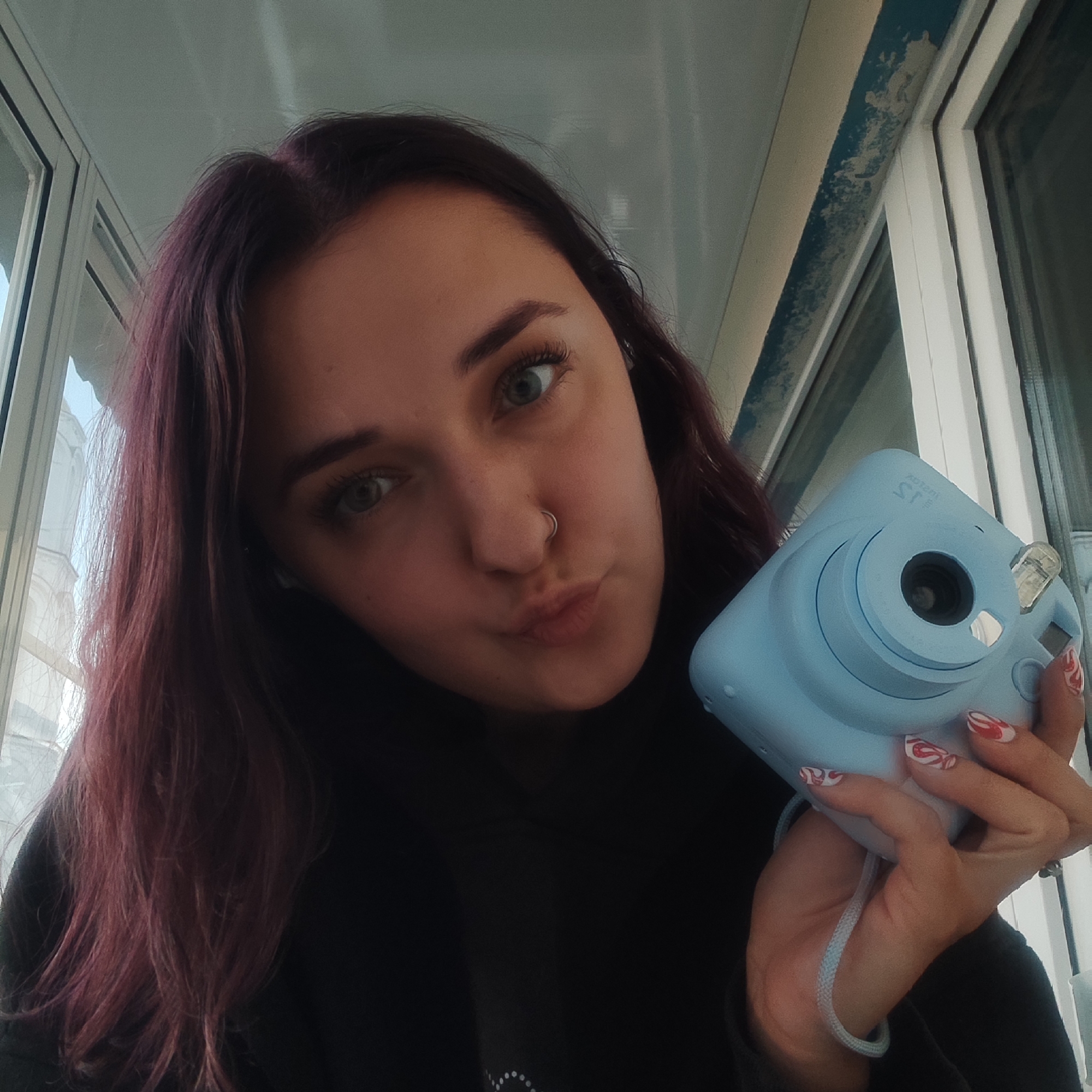 A selfie-style portrait captures a young woman looking directly into the camera with a playful, slightly pouting expression. She has long, dark reddish-purple hair, striking light green eyes, and a silver hoop nose ring on her left nostril. She is wearing a dark, likely black, top.
In her hands, she holds a light blue Fujifilm Instax Mini 11 instant camera. The camera's branding, "instax mini 11," is visible on its front, appearing mirrored due to the selfie angle. Her fingernails are adorned with a distinctive red and white swirling pattern.
The setting appears to be indoors, with natural light streaming in through a large window visible in the background. Outside the window, parts of a white building with architectural details, possibly columns or ornate facades, can be glimpsed, suggesting an urban environment. Above the window, there is a visible strip of light blue paint that shows signs of peeling. The window frames are white. The bright natural light indicates it is daytime. The overall scene depicts a casual, personal moment. The location context provided is Yekaterinburg, Russia.