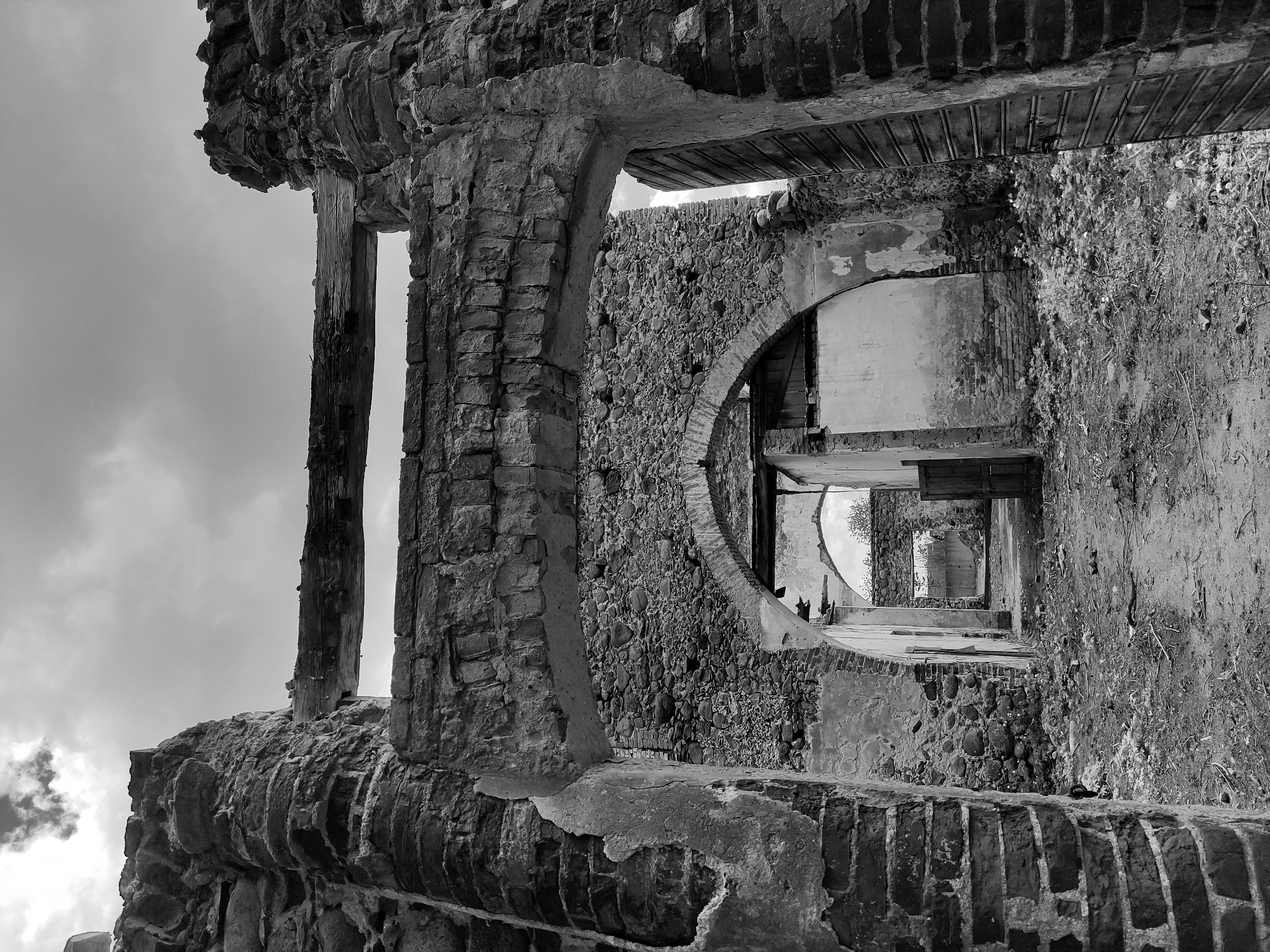 The image shows a partially ruined brick building in black and white. The building's walls are crumbling, revealing the brickwork and stone construction. There are arched openings and doorways leading into the interior of the structure. The sky is visible through openings in the building. The location is Жалудоцкі сельскі Савет, Belarus. The scene is static, depicting the state of disrepair and abandonment of the building.