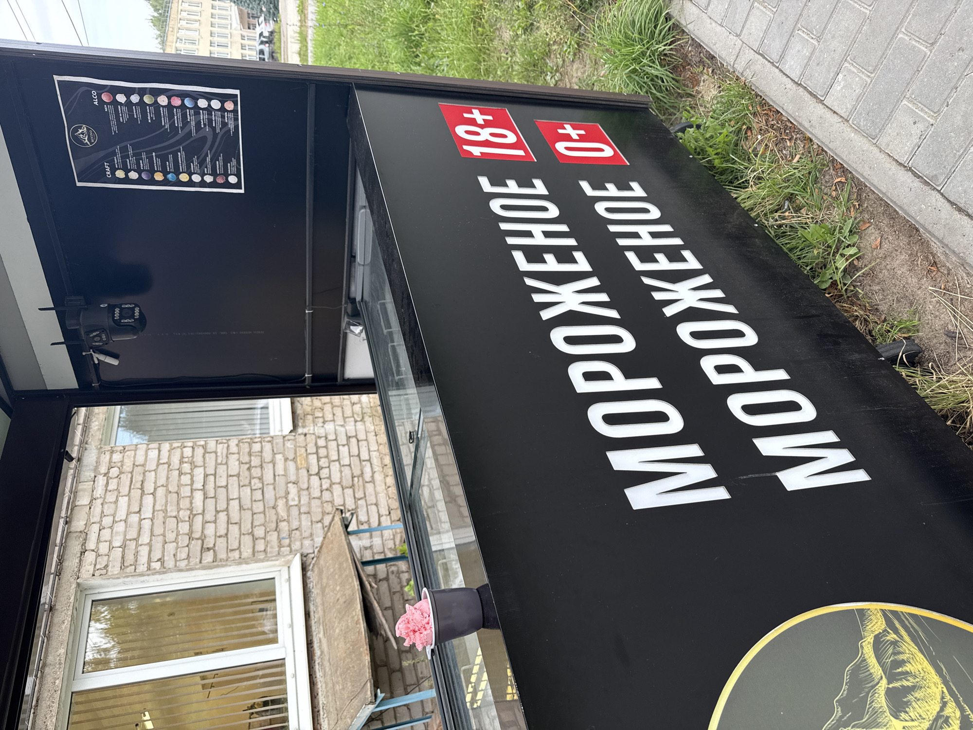 An outdoor ice cream stand is shown in the city of Chaikovsky, Russia. The stand has a black exterior with white lettering that reads "МОРОЖЕНОЕ" (ice cream) twice, with age restrictions "18+" and "0+" displayed in red squares next to the second instance of the word. A grey cup with pink ice cream sits on the edge of the stand. A menu board displaying different ice cream flavors is visible on the upper back wall of the stand, along with two security cameras. The stand is located next to a brick building and a paved sidewalk with grass growing alongside it. The overall scene suggests a commercial establishment offering ice cream.