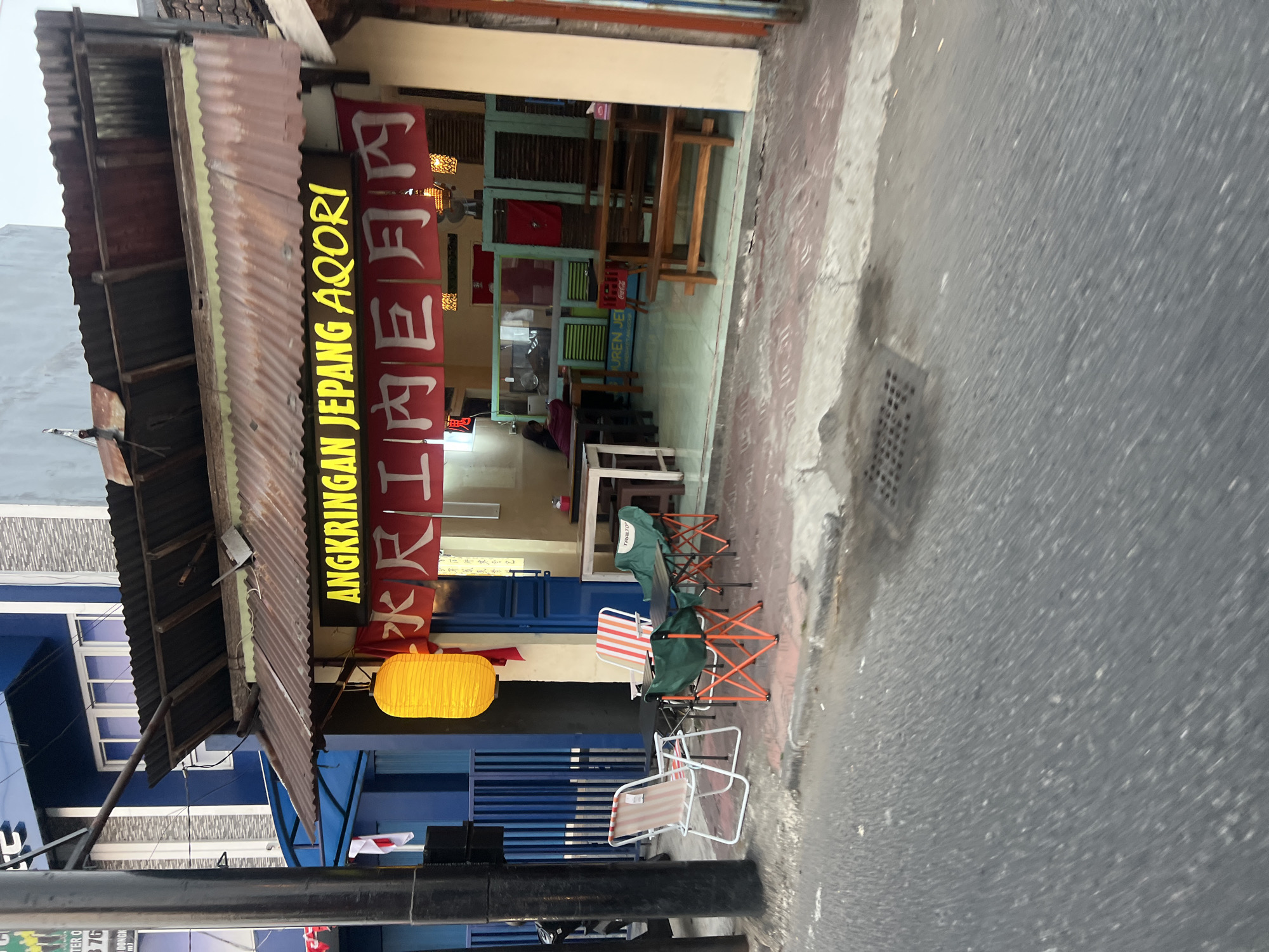 The image depicts a small shop with a rustic corrugated metal roof, identified by the sign "ANGKRINGAN JEPANG AQORI" in yellow neon letters. Below this, red banners with white Japanese characters hang. A bright orange paper lantern is suspended from the awning. In front of the shop, a sidewalk area is set up with portable chairs and tables, including two striped folding chairs, two green camping chairs with orange frames, and a small black table. To the right, there are wooden tables and benches. Through the open doorway, a person is visible sitting inside the shop, which has a counter and what appears to be a kitchen or serving area. The shop is situated on a street, with a road in the foreground and buildings in the background. The overall scene suggests a casual eatery, possibly in a bustling urban area. The presence of Japanese characters and the name "AQORI" could indicate a Japanese-inspired food stall. The setting appears to be in a commercial district, with evidence of street life and pedestrian activity. The image is taken from a slightly elevated perspective, looking towards the shop. The lighting suggests it is either early morning or late afternoon.