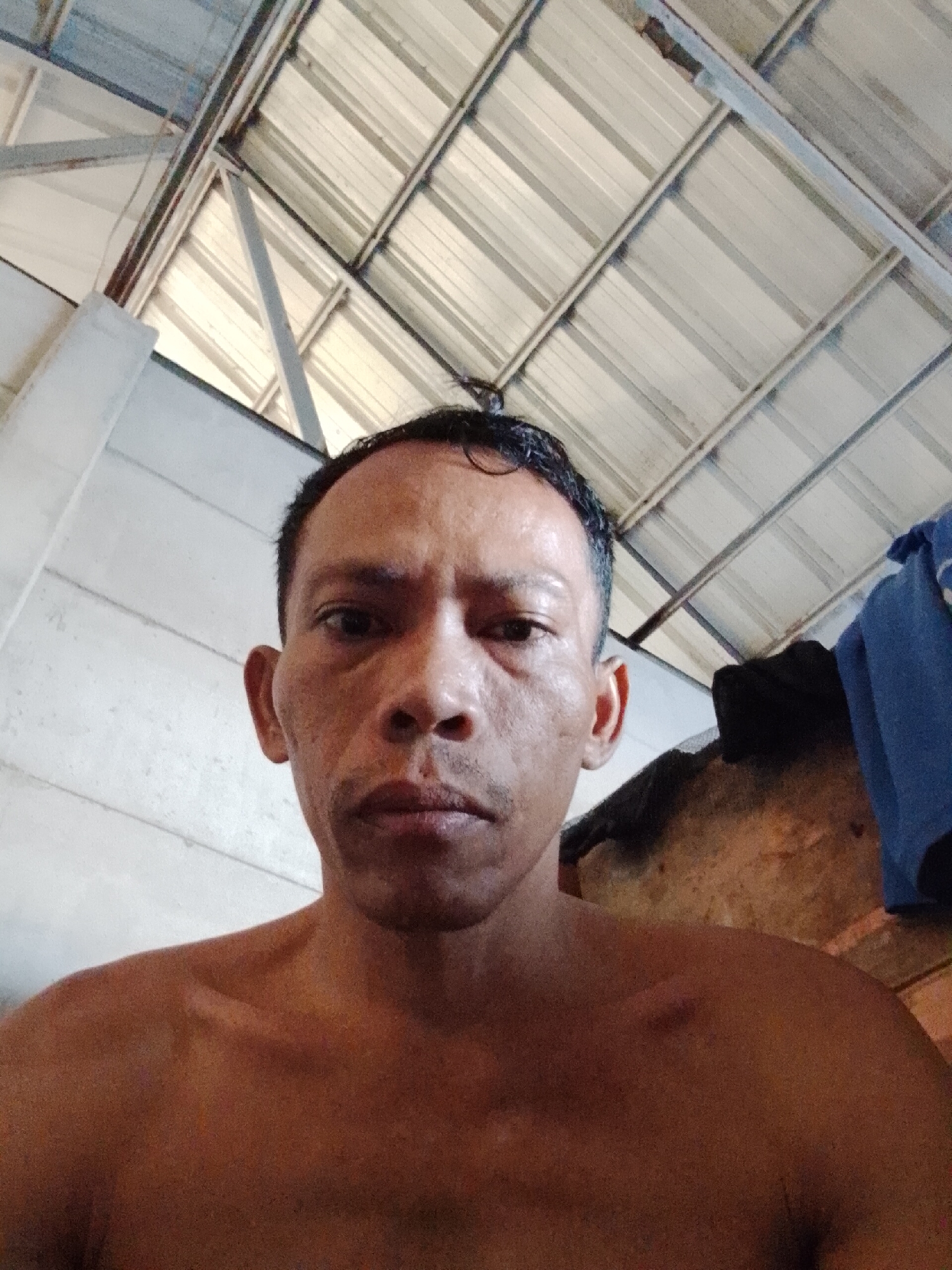 A shirtless man is indoors, looking into the camera. The location is likely Bukit Ketok, Indonesia, based on metadata. The background includes a corrugated metal roof, possibly of a simple dwelling, and a wall. A blue garment is draped near the background's right side.