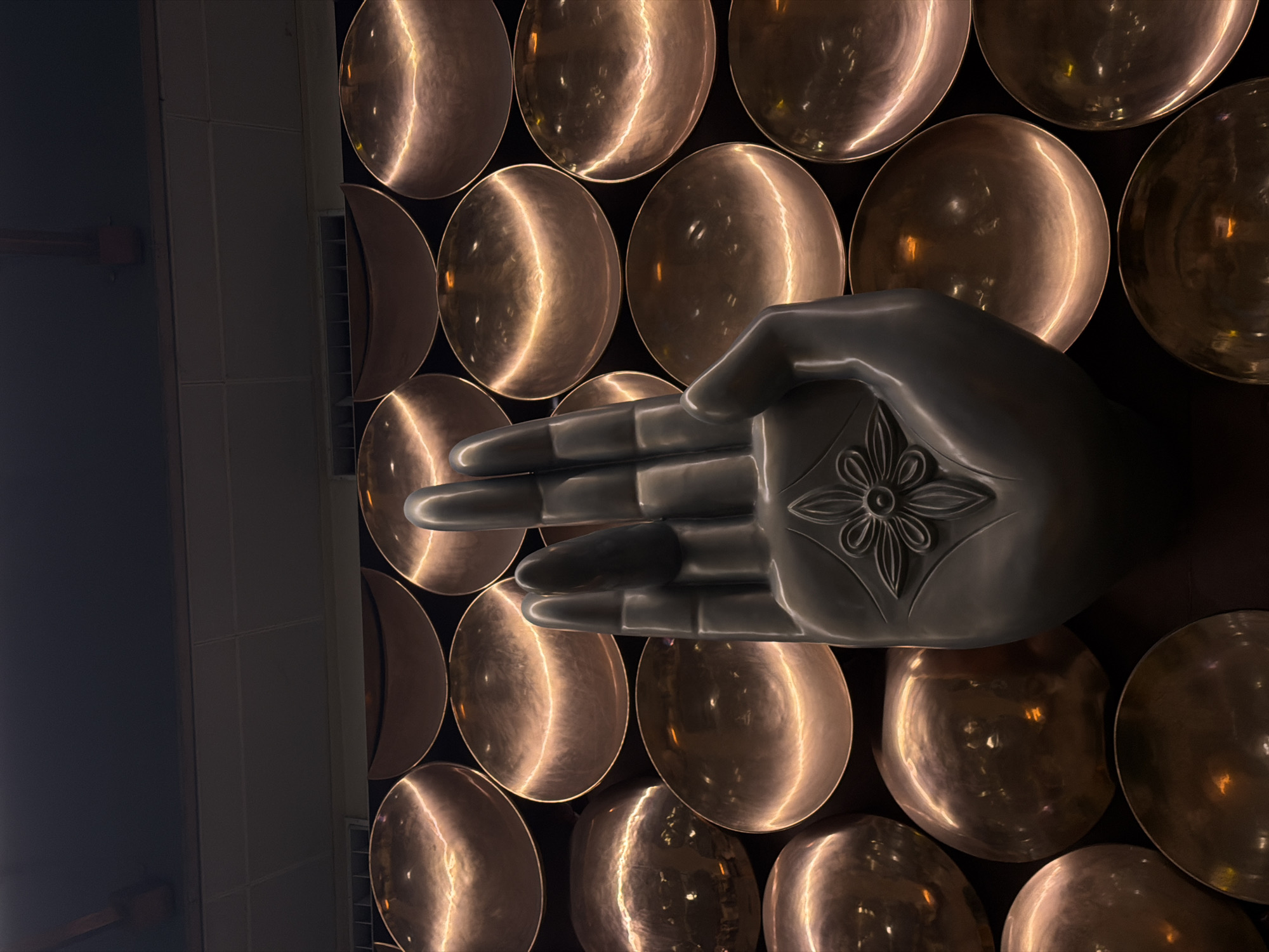 The image features a large, metallic sculpture of a hand with an open palm, adorned with a floral motif. The sculpture is positioned in front of a wall covered in a grid of large, circular, concave metallic objects that reflect light, creating a warm, coppery glow. The overall aesthetic is one of modern, abstract art with a spiritual or meditative undertone. The setting appears to be indoors, possibly in a public space or an art installation. Given the style and the presence of such sculptures, a likely location in Delhi, India, would be an airport terminal or a contemporary art gallery. The lighting is dim, with spotlights highlighting the reflective surfaces.