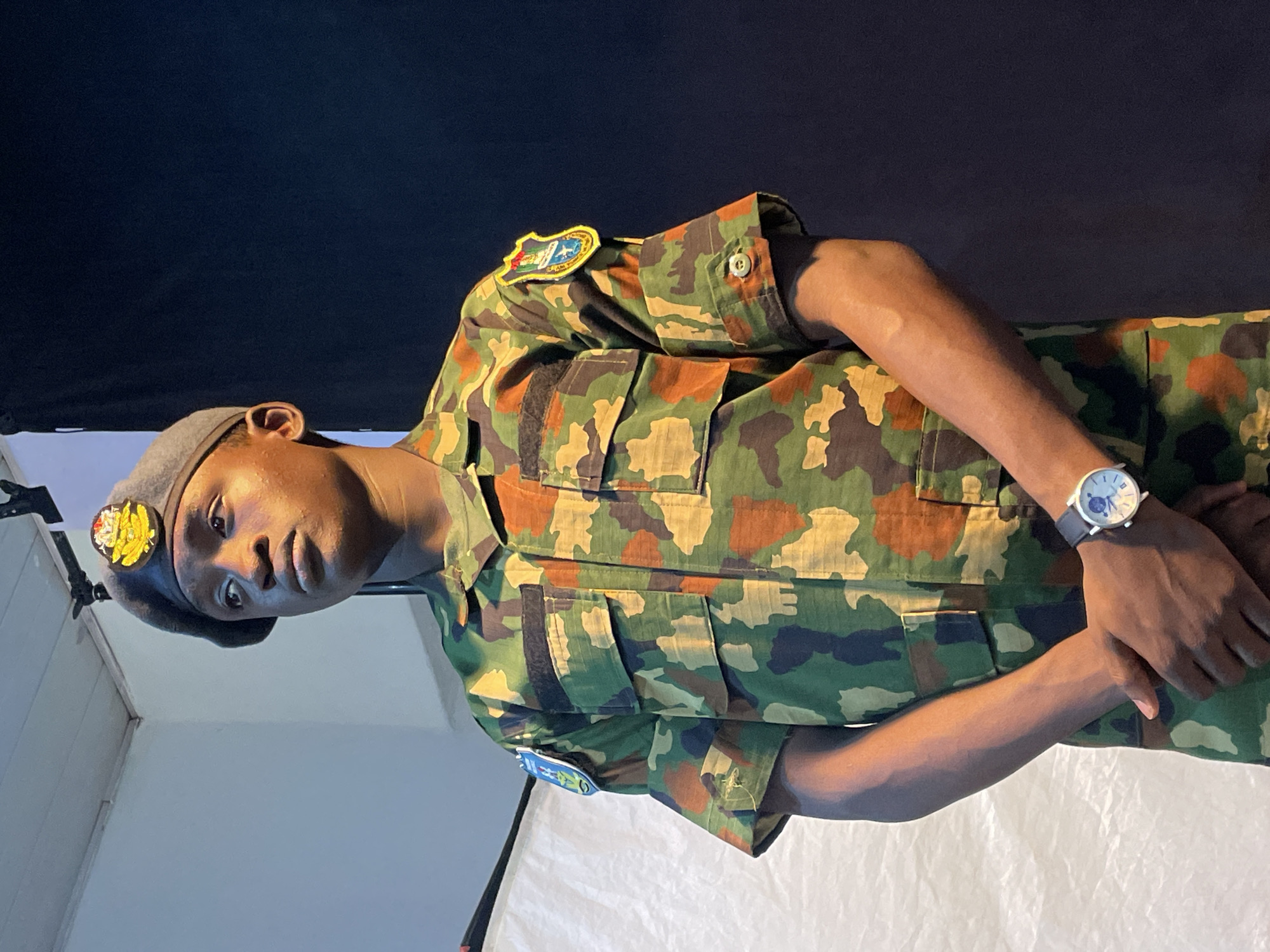 A person wearing a camouflage uniform and a gray beret with a badge is visible in this photograph. The individual has dark skin and is wearing a wristwatch on the left wrist. Patches are affixed to the upper sleeves of the uniform. The background is a combination of white and black. The location is not discernible as being Barakin Akawo, Nigeria.