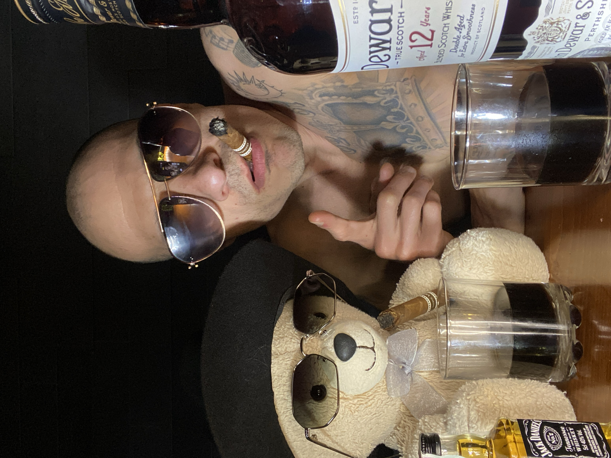 The image features a shirtless man with tattoos, wearing sunglasses and smoking a cigar. He appears to be lying down, with a bottle of Dewar's 12-year-old Scotch whiskey positioned above him. Next to him is a glass filled with a dark liquid. Below him, a teddy bear wearing sunglasses and a hat also has a cigar and a glass of a dark liquid. A small bottle of Jack Daniel's whiskey is also visible. The location, Pleven, Bulgaria, is not evident from the image itself. The background is dark and unclear, obscuring any environmental details.