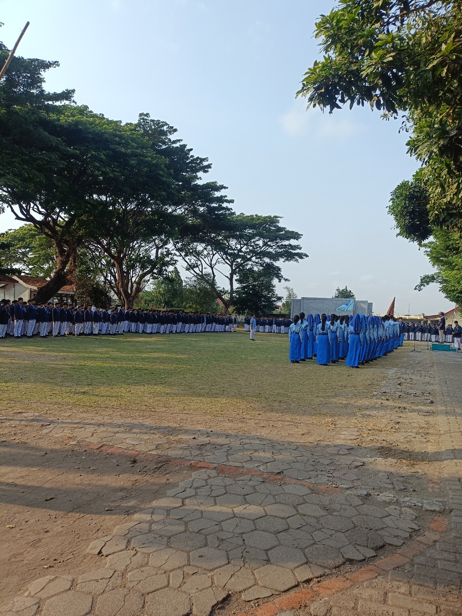 An outdoor scene in Kediri City, Indonesia, features a large group of uniformed students standing in formation on a grassy field. The students, appearing to be high school-aged, are predominantly wearing blue uniforms, with some in white shirts and dark trousers. They are arranged in multiple rows, facing a central area where other students are also gathered. A light blue flag is visible, held by one of the students in blue. In the background, there are buildings and large trees, suggesting the location is likely a school campus or a public park. The ground in the foreground is a paved area with hexagonal stone tiles. The sky is clear and blue, indicating it is daytime. The overall impression is of an organized outdoor gathering or ceremony.