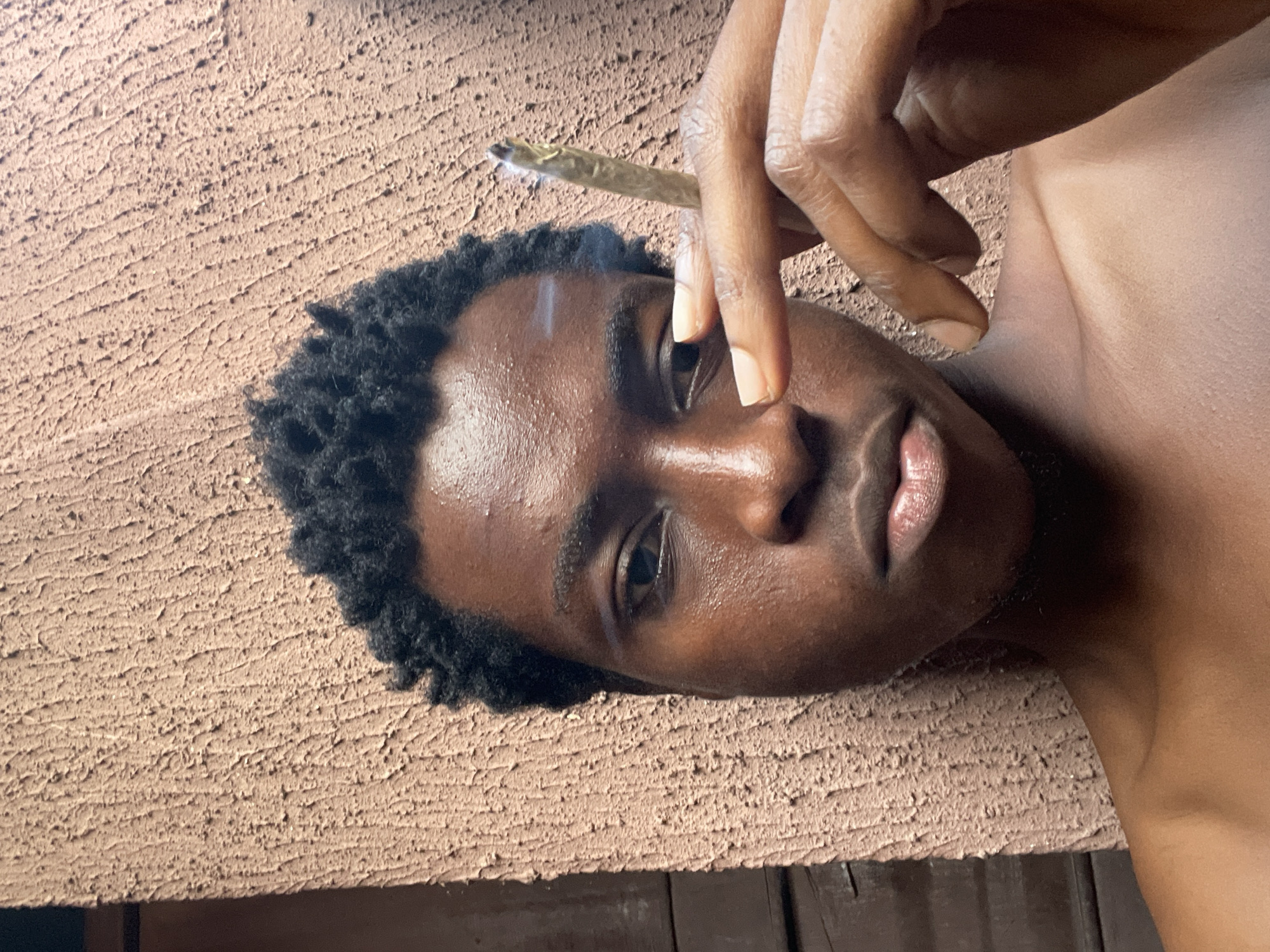 The image shows a close-up, horizontally oriented shot of a person's face and upper body. The individual appears to be lying down, with their head resting on a textured, brown surface that resembles a wall or rough plaster. Their skin is dark and appears slightly moist, reflecting the light. They have short, dark, curly hair.
The person is holding a lit cigarette or joint between their index and middle fingers, bringing it towards their mouth. A wisp of smoke is visible rising from the end of the cigarette, just above their forehead. Their eyes are open and looking towards the camera with a neutral expression. Their lips are slightly parted. The background behind the person's head and shoulder is a pale, smooth surface, likely their skin.
The lighting suggests it is daytime, possibly late afternoon due to the warm tones. The texture of the surface the person is resting on is prominent in the foreground, with visible ridges and indentations. The overall scene conveys a moment of relaxation or introspection. No visible text is present in the image. The location context of Nkpor, Nigeria, suggests an outdoor or possibly indoor setting with elements common to that region.