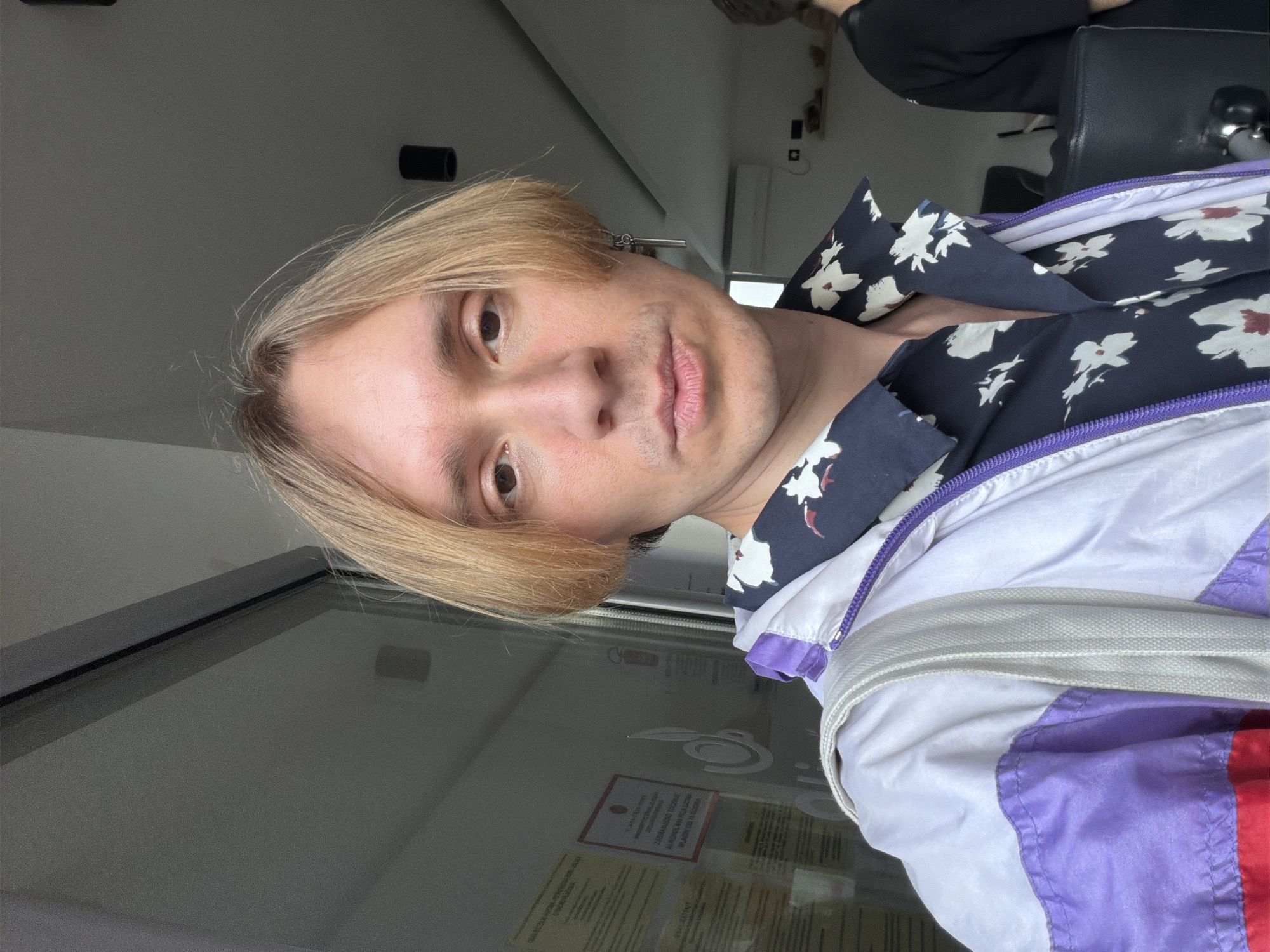 The image features a young person, centered and looking directly at the camera, taking a selfie indoors in what appears to be a public or commercial establishment in Budva, Montenegro. The individual has blonde, shoulder-length hair with a slight side part and light stubble on their chin and upper lip. Their expression is neutral, with lips slightly pursed.
They are wearing a dark blue shirt with a white floral pattern, possibly depicting hibiscus flowers with red centers. Over this, they wear a multi-colored windbreaker or jacket, primarily white, with distinct sections of purple and red, and a visible purple zipper.
The background includes a light grey ceiling with black light fixtures. To the right, another person is partially visible, seated in a dark chair. A prominent feature in the lower background is a reflective surface, likely a glass partition or counter, behind which several documents are displayed. One notable red-bordered paper clearly shows the text "ZABRANJENO FOTOGRAFSANJE", translating to "PHOTOGRAPHY FORBIDDEN", while another below it has "INFORMACIJE" ("INFORMATION"). The overall lighting suggests it is daytime, though no direct sunlight is evident.