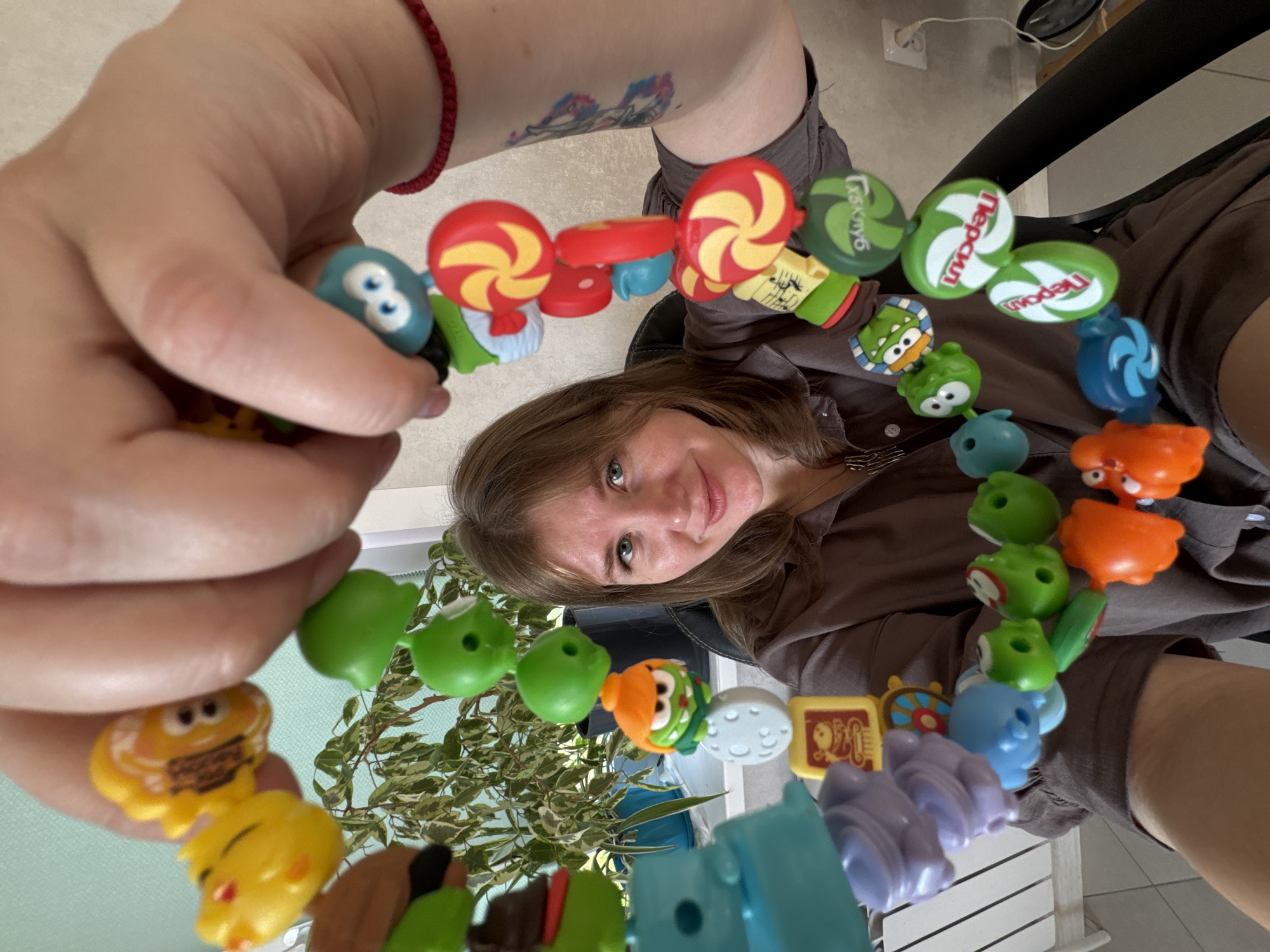 A woman holds a toy chain, possibly from the "Angry Birds" franchise, towards the camera. The setting appears to be inside a building in Ферма, Russia. The toy chain consists of various character heads and other shapes in different colors. A small potted plant is partially visible in the background, adding to the indoor setting. The woman is sitting, wearing a brown shirt, with a portion of a black chair visible behind her. A tattoo can be seen on the woman's arm.