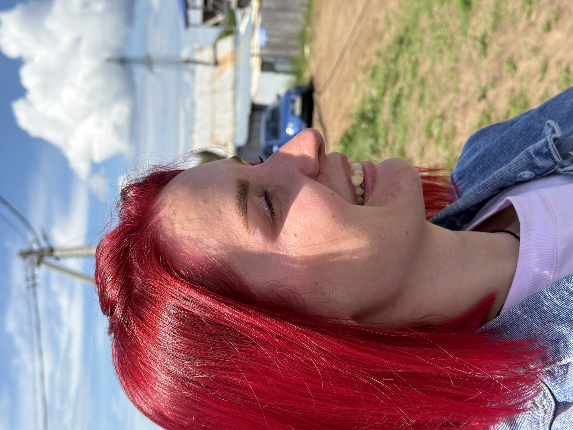 The image shows a woman with bright red hair, smiling with her eyes closed. She is wearing a white top under a denim jacket. The background includes a blue sky with white clouds, power lines, a building with a corrugated metal roof, a glimpse of a blue car, and some green vegetation. It is likely an outdoor setting on a sunny day. The exact location in Chaikovsky, Russia, cannot be determined from the image.