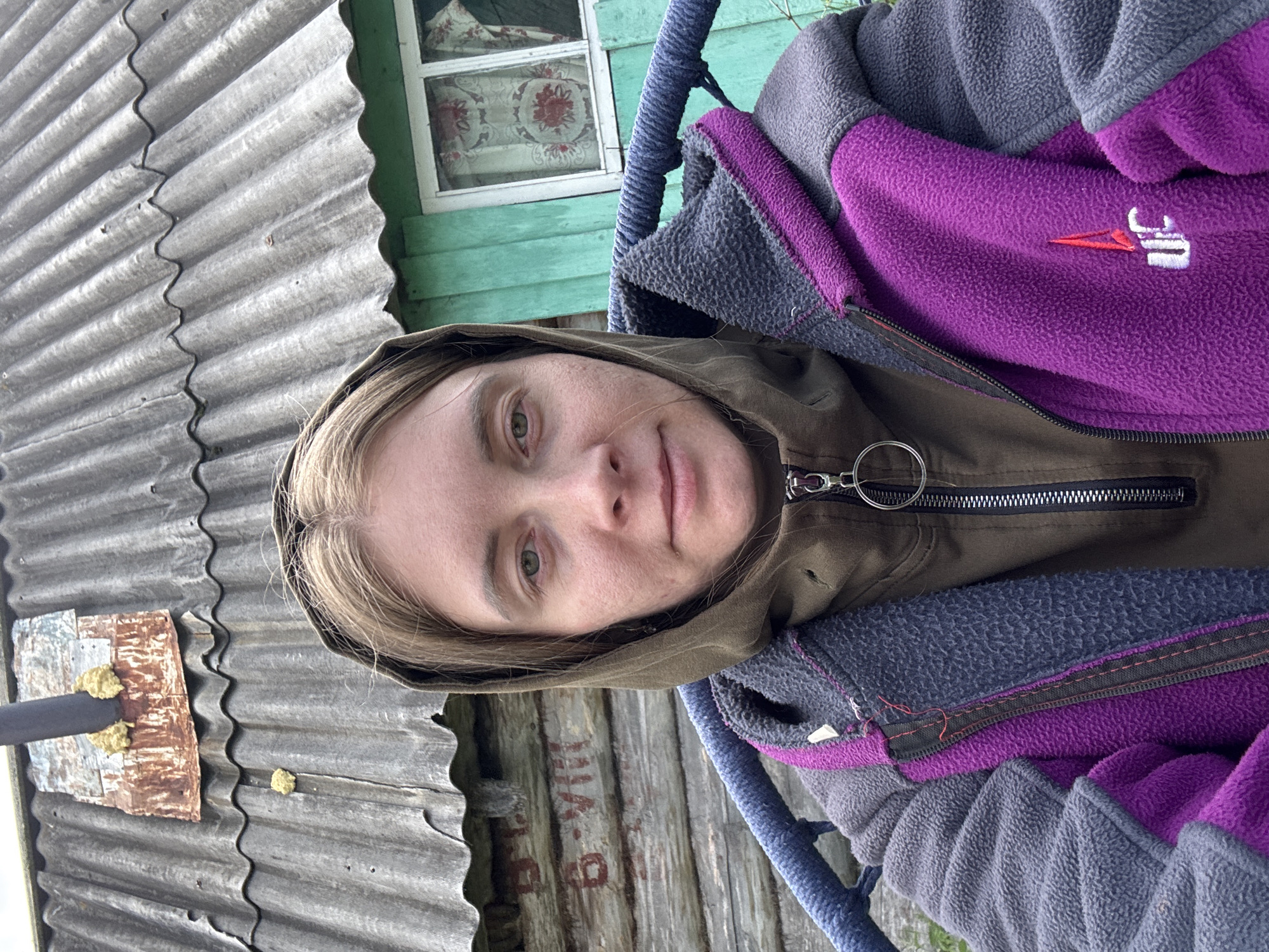 A medium shot captures a young Caucasian woman with light brown hair and fair skin, wearing a brown hooded sweatshirt and a purple and gray fleece jacket. She is seated in a chair with a blue rope-like armrest, facing the camera. Her gaze is directed towards the viewer. Behind her, a weathered, gray, corrugated roof covers a wooden building with green trim and a window with floral curtains. A rusty metal plate with yellow foam insulation is affixed to the roof near a dark pipe. Red lettering is visible on the wooden wall of the building. The scene suggests an outdoor setting in a rural or semi-rural area, possibly on the outskirts of the city of Курган, Russia, given the architectural style and materials.