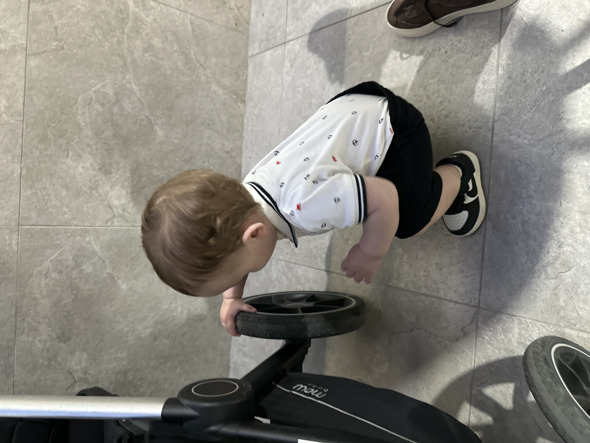 A young child is crouching next to a stroller on a tiled floor. The child is wearing a white polo shirt with a pattern of small red and black anchors, and black shorts. They are also wearing black and white sneakers. The stroller is black with a silver handle and a wheel visible. The floor is tiled with large, grey, stone-like tiles. The background shows a light-colored tiled wall. The lighting suggests an indoor environment. There is a shadow of the child and stroller cast on the floor. A brown shoe is visible to the right of the frame. No specific landmarks indicating the city of Chișinău, Moldova, are visible in the image.