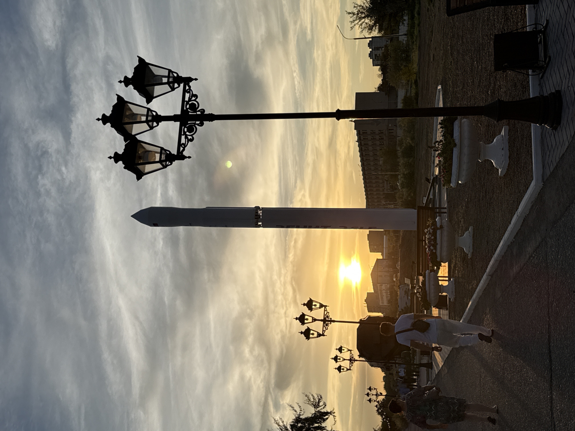 The image captures a sunset scene in the city of Baikonur, Kazakhstan, with a prominent rocket monument as the central subject. The sky is filled with dramatic clouds, illuminated by the low sun, creating a warm, orange and yellow glow. A multi-headed lamppost, a characteristic feature of Baikonur's streetlights, stands in the foreground, silhouetted against the sky.
In the middle ground, two people are walking away from the camera on a paved pathway. The person in front is wearing a white t-shirt and light-colored pants, with a small bag slung across their chest. The person behind them is wearing a patterned dress and carrying a bag. To the left of the pathway, more lampposts and a line of trees are visible.
Behind the rocket monument, a large, multi-story building with rows of windows can be seen, suggesting an urban or residential area. The ground around the monument is a mix of gravel and dark soil, with decorative flower planters. The overall atmosphere is one of a serene evening walk, with the iconic Baikonur rocket serving as a backdrop. The lighting suggests it is late afternoon or early evening, as the sun is setting.