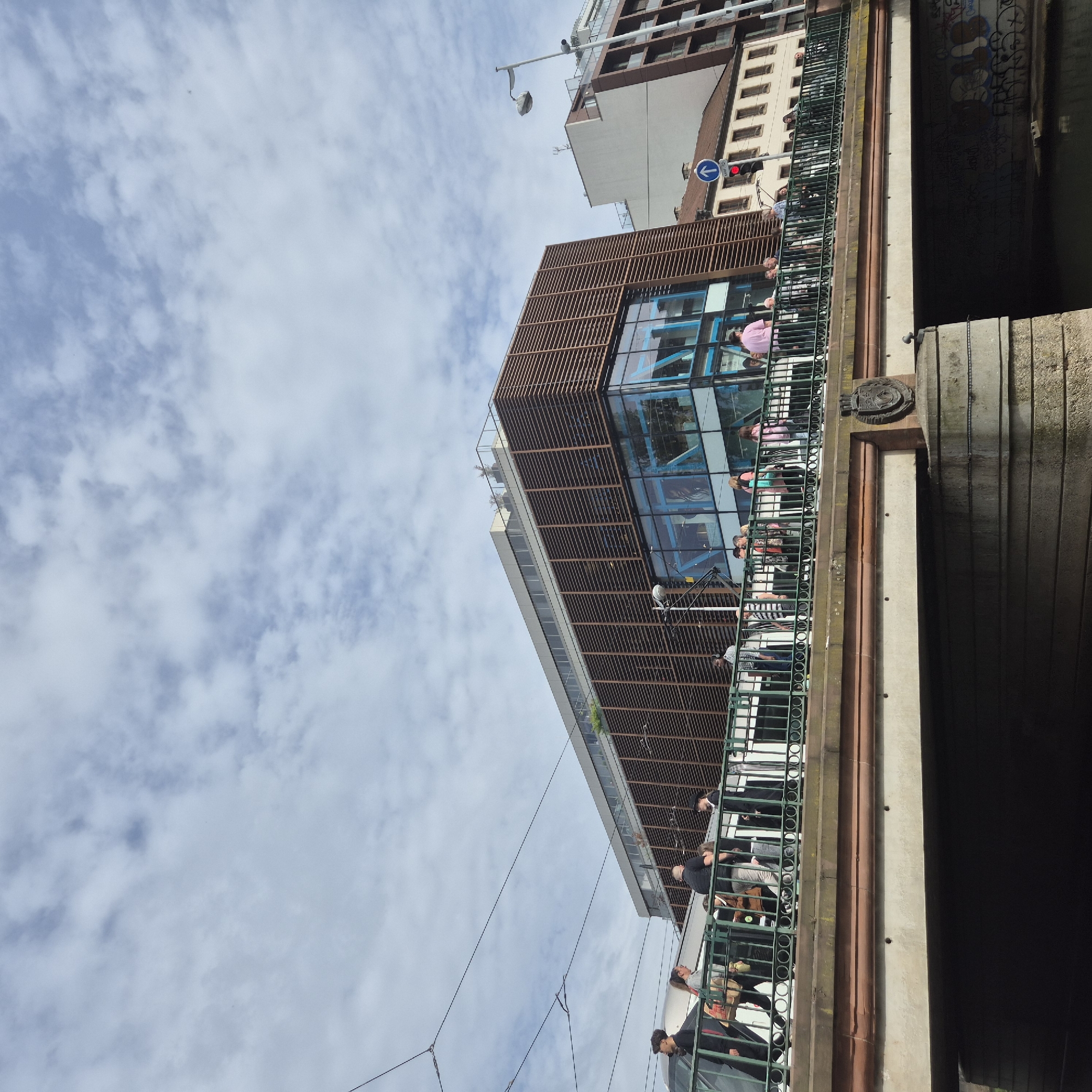 The image is a low-angle shot taken from beneath a bridge in Strasbourg, France, capturing a partial view of a modern building with a brown, slatted facade and glass windows. The building is situated adjacent to the bridge and appears to have multiple levels with people visible inside. A pedestrian bridge runs alongside the building, with people walking along it. Overhead tram lines stretch into the distance.
To the right, a section of the bridge is visible, constructed with concrete and brown trim. The underside of the bridge is dark, creating a strong contrast with the sky and building. The sky is partly cloudy with patches of blue visible.
Other buildings are seen in the background, indicating an urban environment. A street light and a traffic signal are also visible further back. Overall, the scene depicts a mix of modern architecture and urban infrastructure against a cloudy sky.