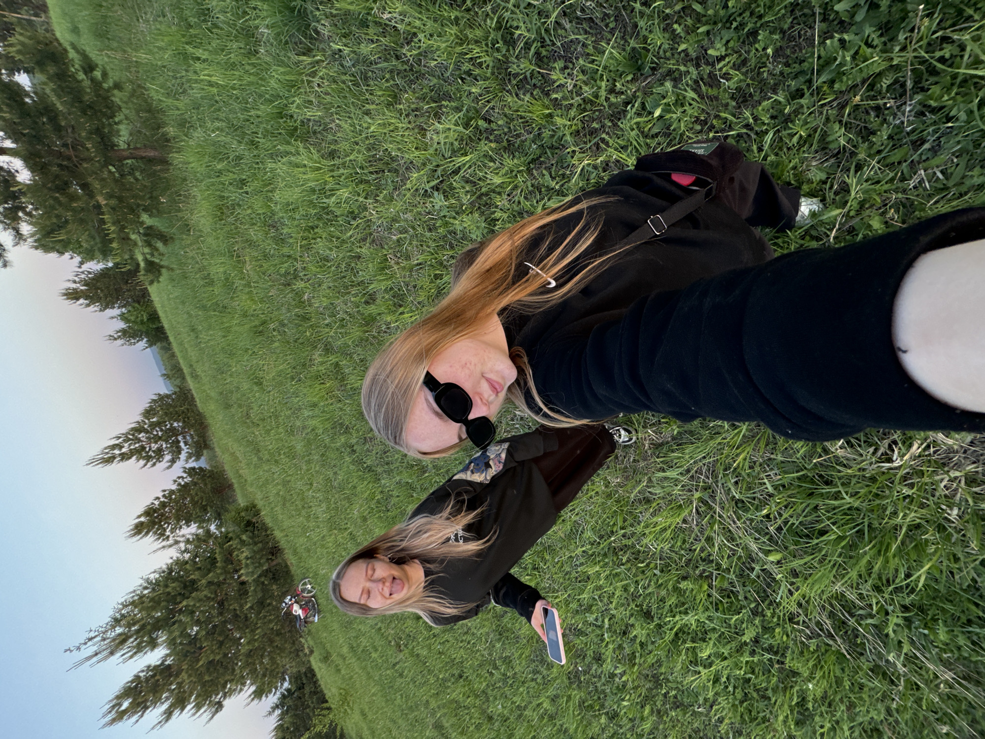 Two young women are taking a selfie in a grassy field, possibly Zhigalki, Russia. One woman wears sunglasses and a black top. The other woman holds a cell phone. A bicycle rests near trees in the background.