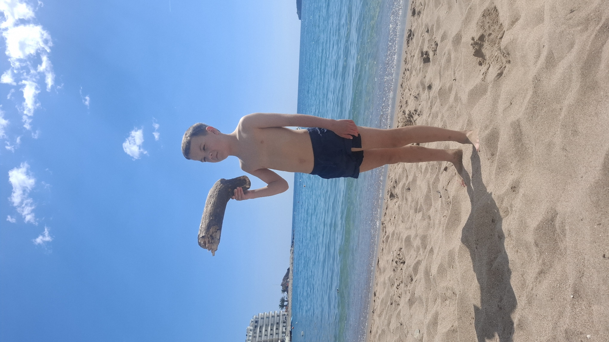 The image shows a shirtless young boy in dark blue swim trunks standing on a sandy beach next to the shoreline of the Adriatic Sea. He is holding a piece of driftwood in his left hand. The sky is bright blue with scattered clouds. Behind the boy is the sea, with a building visible in the distance. The beach is light brown sand. The image appears to be taken near Budva, Montenegro, based on the presence of buildings and beaches common to that coastal city.