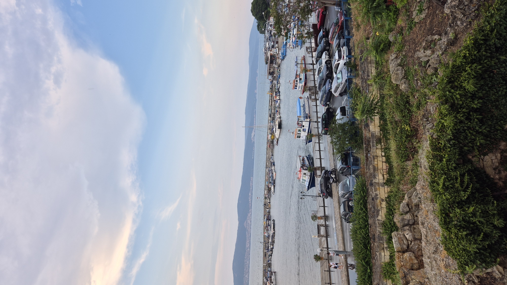 The image shows a view of the Nesebar harbor in Bulgaria. In the foreground, there is a rocky embankment covered in green ivy, with some larger rocks scattered among the vegetation. Behind this, a paved area borders the harbor. Several cars are parked along this paved area, including a black sedan, a black motorcycle, a silver SUV, and various other vehicles. Two people are walking on the sidewalk near the parked cars, and a streetlamp stands on the pavement.
The harbor itself is filled with numerous boats of various sizes. Some are docked along piers, while others are moored in the water. Two passenger boats are prominently featured in the middle ground, one of which appears to be carrying passengers. Further out in the harbor, more boats are visible, including a sailboat with a tall mast.
Beyond the harbor, a body of water stretches to the horizon, with a hilly coastline visible in the distance. The sky above is a pale blue with scattered white and wispy clouds, tinged with a soft yellow and orange hue, suggesting either dawn or dusk. The overall impression is a calm, coastal scene in a historic Bulgarian town.
