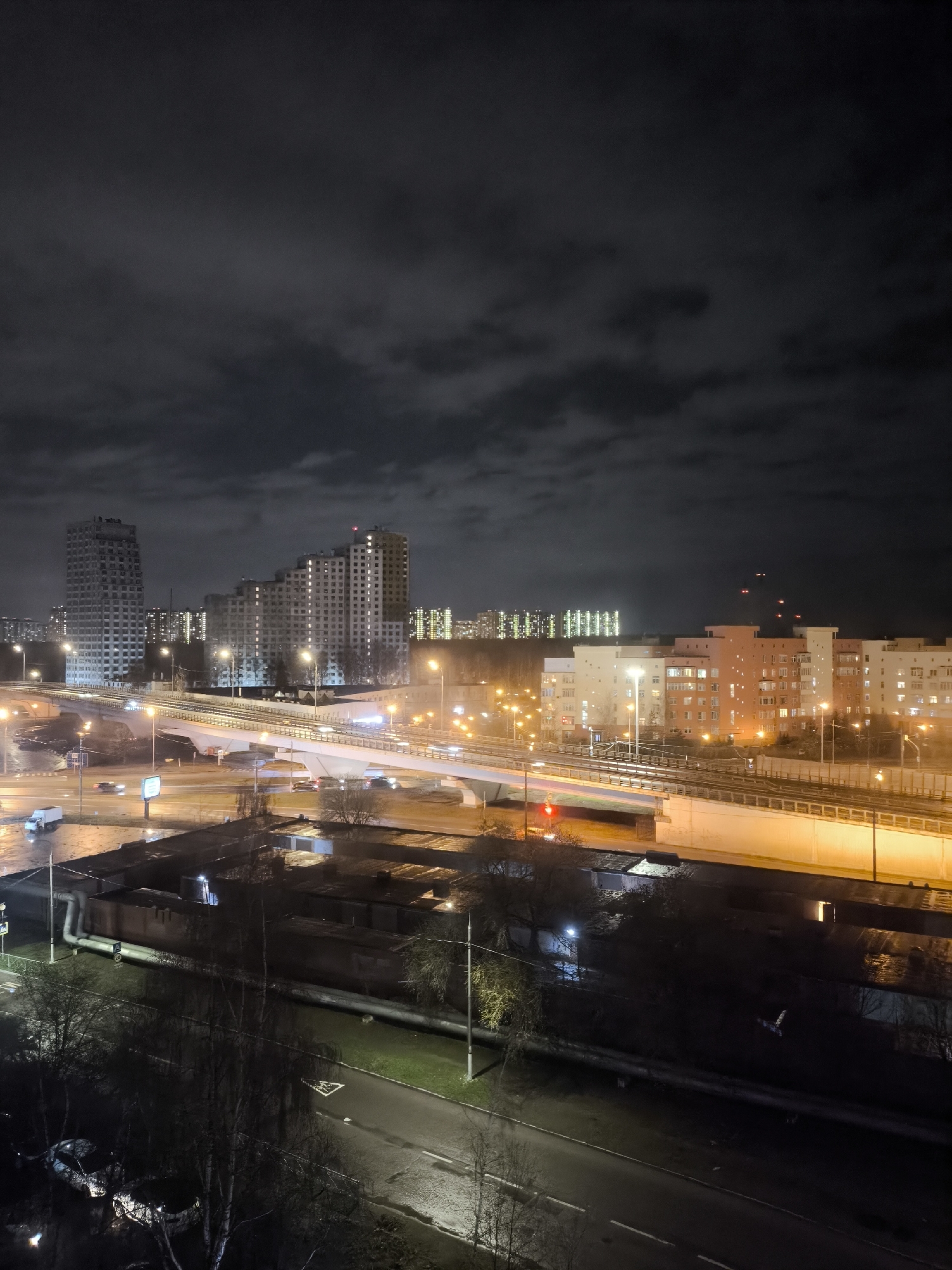 The image presents a nighttime cityscape in Moscow, Russia, under an overcast sky. Several apartment buildings are visible, illuminated with interior lights. A highway overpass, brightly lit with street lamps, spans the lower portion of the image, with traffic visible on the roadways. Below the highway, a lower level of industrial or commercial buildings is present. The foreground depicts a street with wet pavement reflecting the surrounding lights. Trees, appearing bare due to the season, line the street. There is a car parked near the lower left corner of the frame.