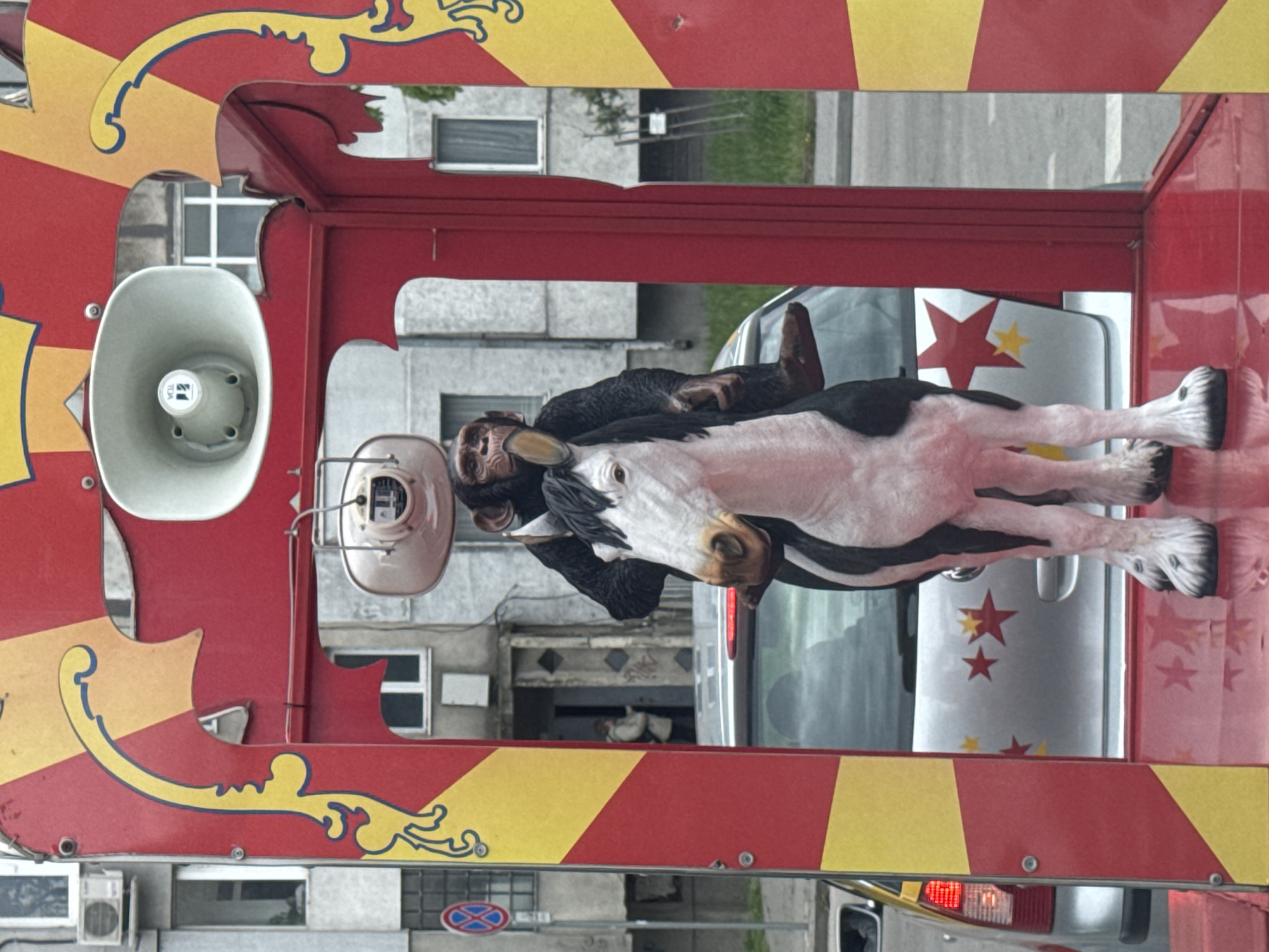 A red and yellow decorated vehicle with a loudspeaker and a monkey and horse figure is situated on a street in Varna, Bulgaria. The vehicle's design incorporates red and yellow stripes and decorative gold flourishes. Visible outside the vehicle is a gray multi-story building with windows, a pedestrian crossing sign, and passing vehicles. Two people are visible in the background, standing in the doorway of a building.