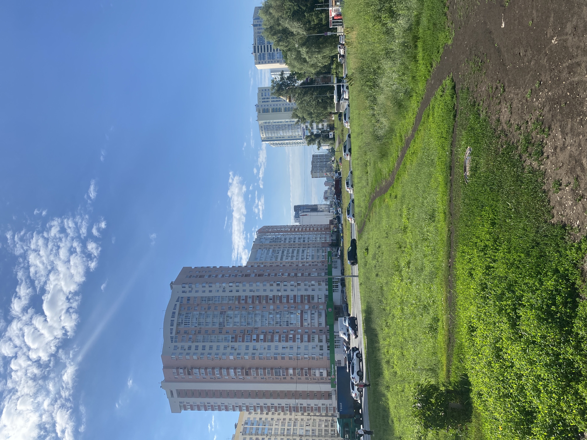 The image depicts a street scene in Novokuybyshevsk, Russia, on a sunny day with scattered clouds. In the foreground, a grassy area with a dirt path leading through it occupies the lower portion of the frame. To the left, a small, bushy plant is visible. In the mid-ground, a multi-lane road runs horizontally, with several cars and a large green truck driving on it. People can be seen standing on the side of the road. Several tall apartment buildings, characteristic of urban development, line the street in the background. These buildings vary in height and architectural style, with some featuring pink and white facades, while others are lighter in color. Trees are interspersed between the buildings and along the roadside, suggesting a mix of residential and green spaces. The sky is a clear blue with fluffy white clouds. The overall scene conveys a typical urban environment with residential buildings, traffic, and green spaces.